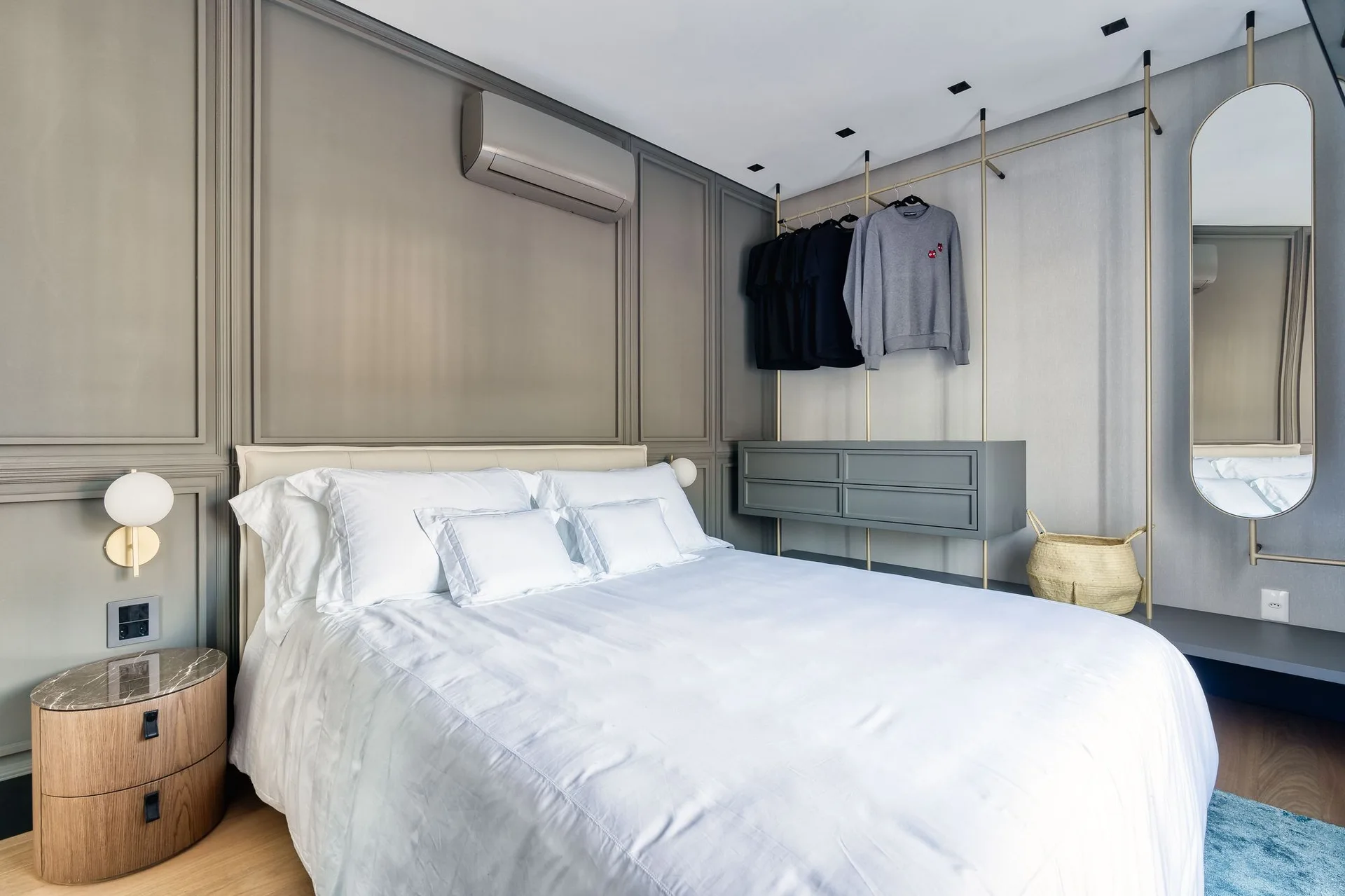 A modern bedroom features a bed with white bedding, a grey paneled wall, a curved wooden nightstand, and an open wardrobe system.