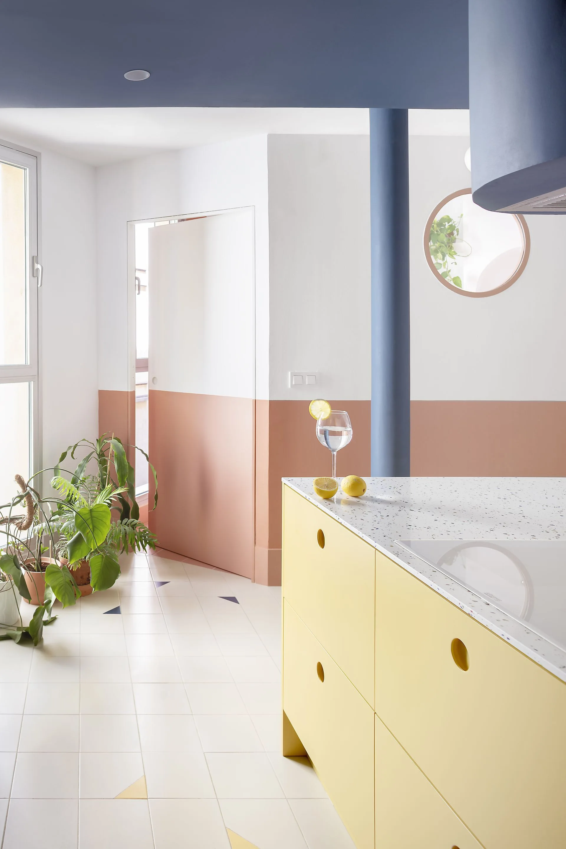 A modern kitchen features a yellow island with a terrazzo countertop, set against color-blocked walls with a blue ceiling and column.