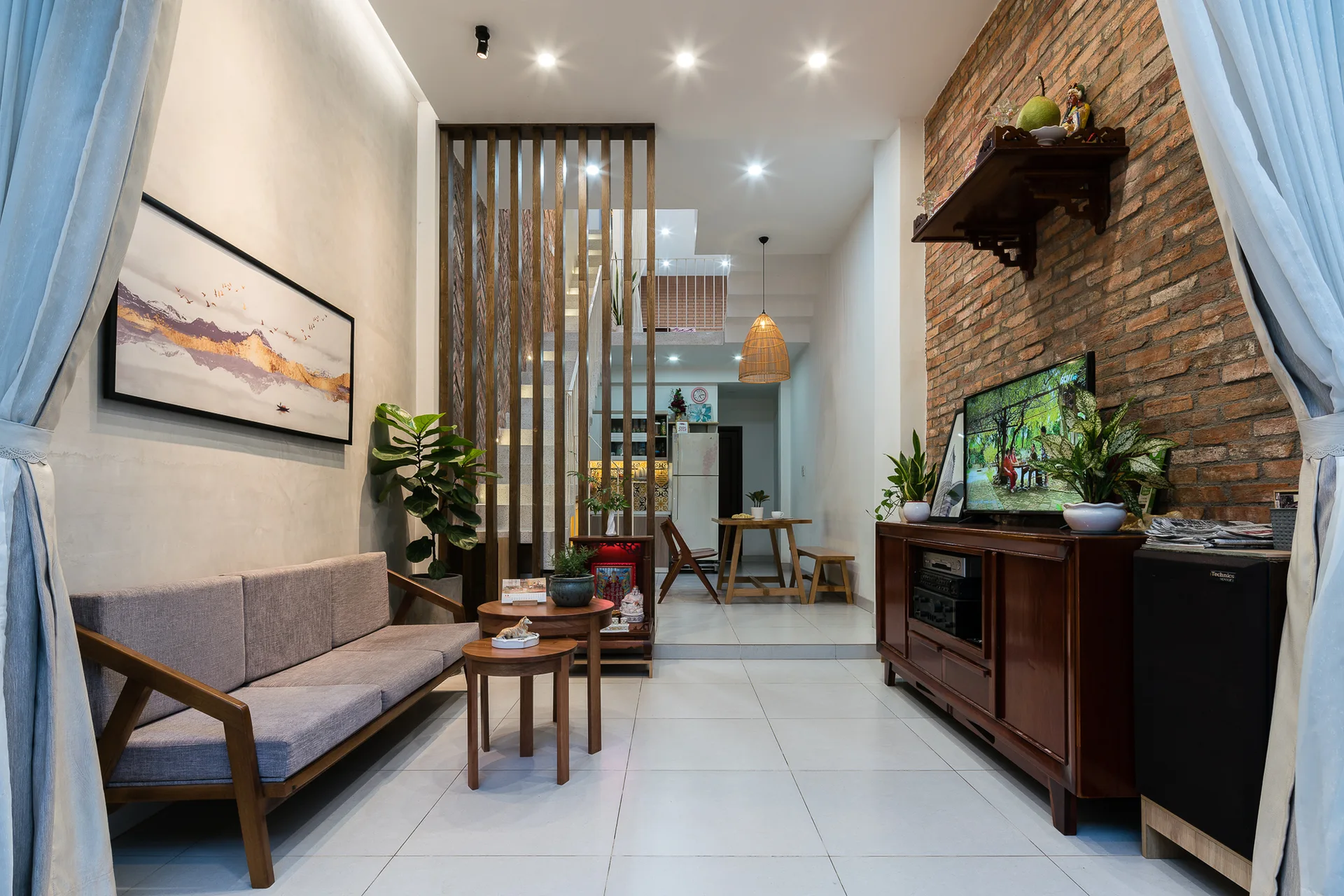 A modern living room features a grey sofa, stacked wooden coffee tables, an exposed brick wall, and a vertical wood slat room divider.