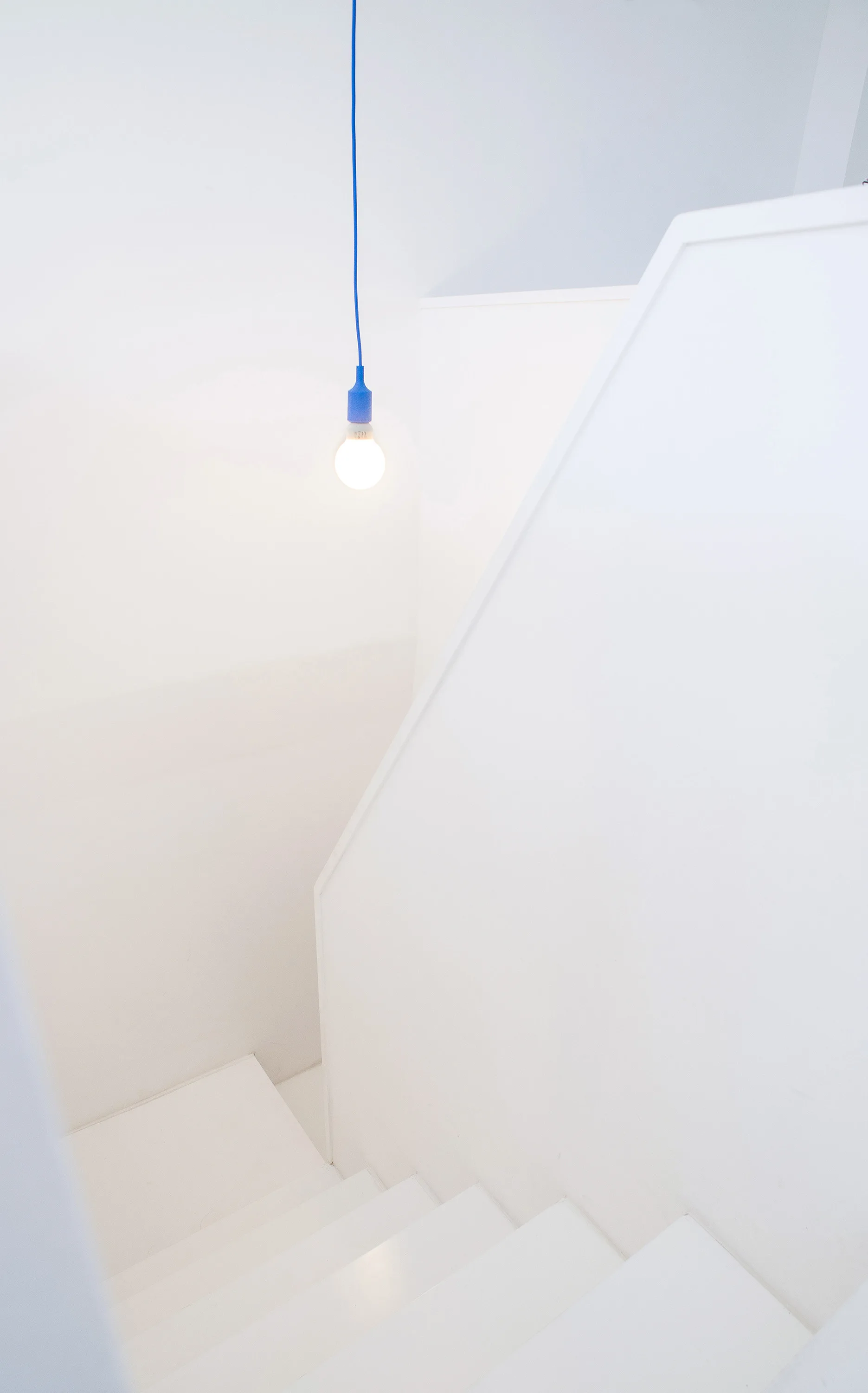 A minimalist white staircase leading downward with an angular banister and a single glowing pendant light hanging from a bright blue cord.