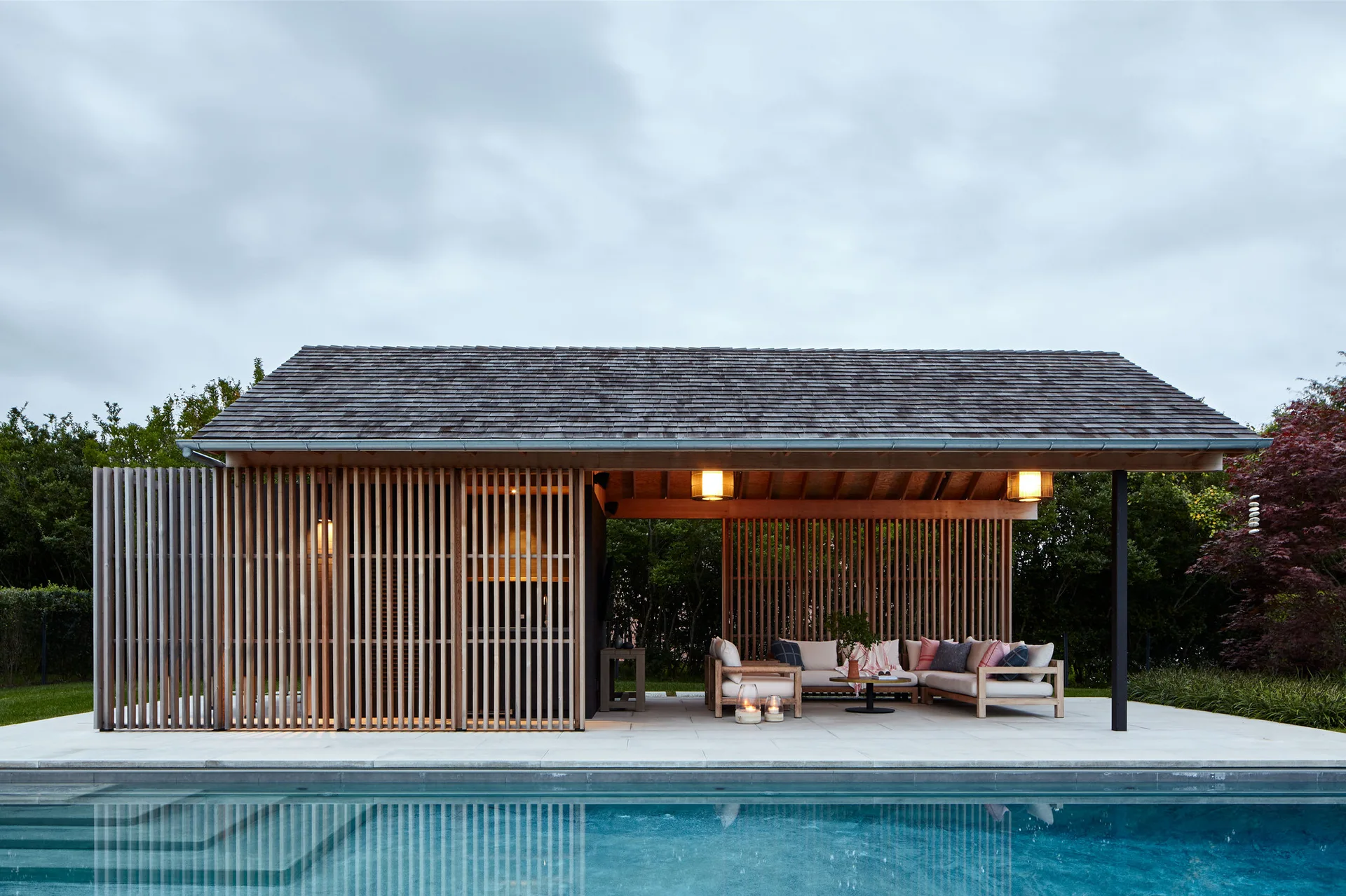 A wood-shingled patio pavilion with vertical timber slat walls features an outdoor sectional sofa next to a swimming pool.