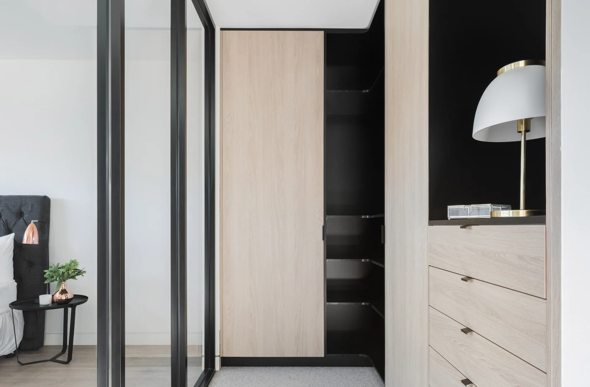 A built-in light wood wardrobe features a matte black interior and integrated drawers, positioned next to a black-framed glass partition.