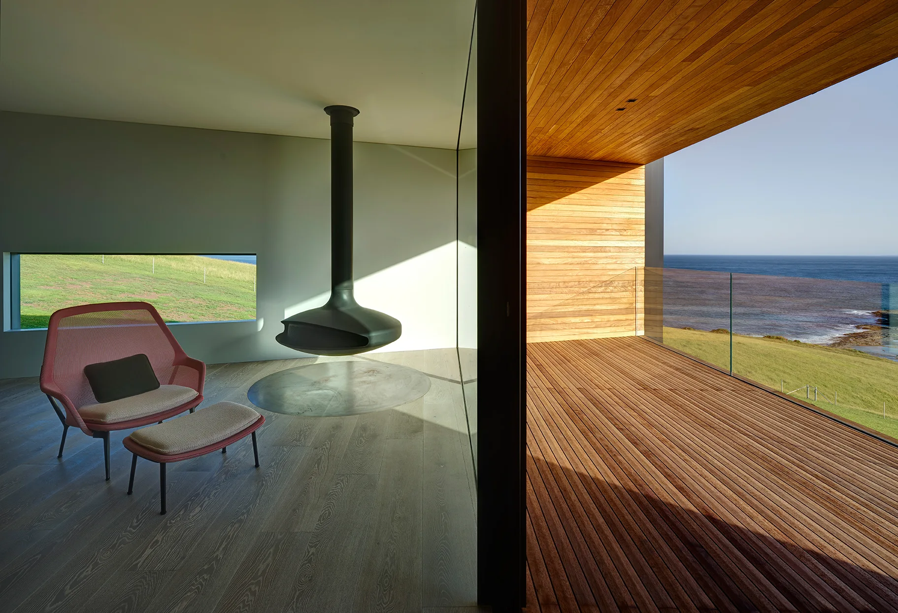 A minimalist indoor lounge features a pink mesh armchair and a black suspended fireplace next to a large glass wall opening onto a wooden coastal deck.