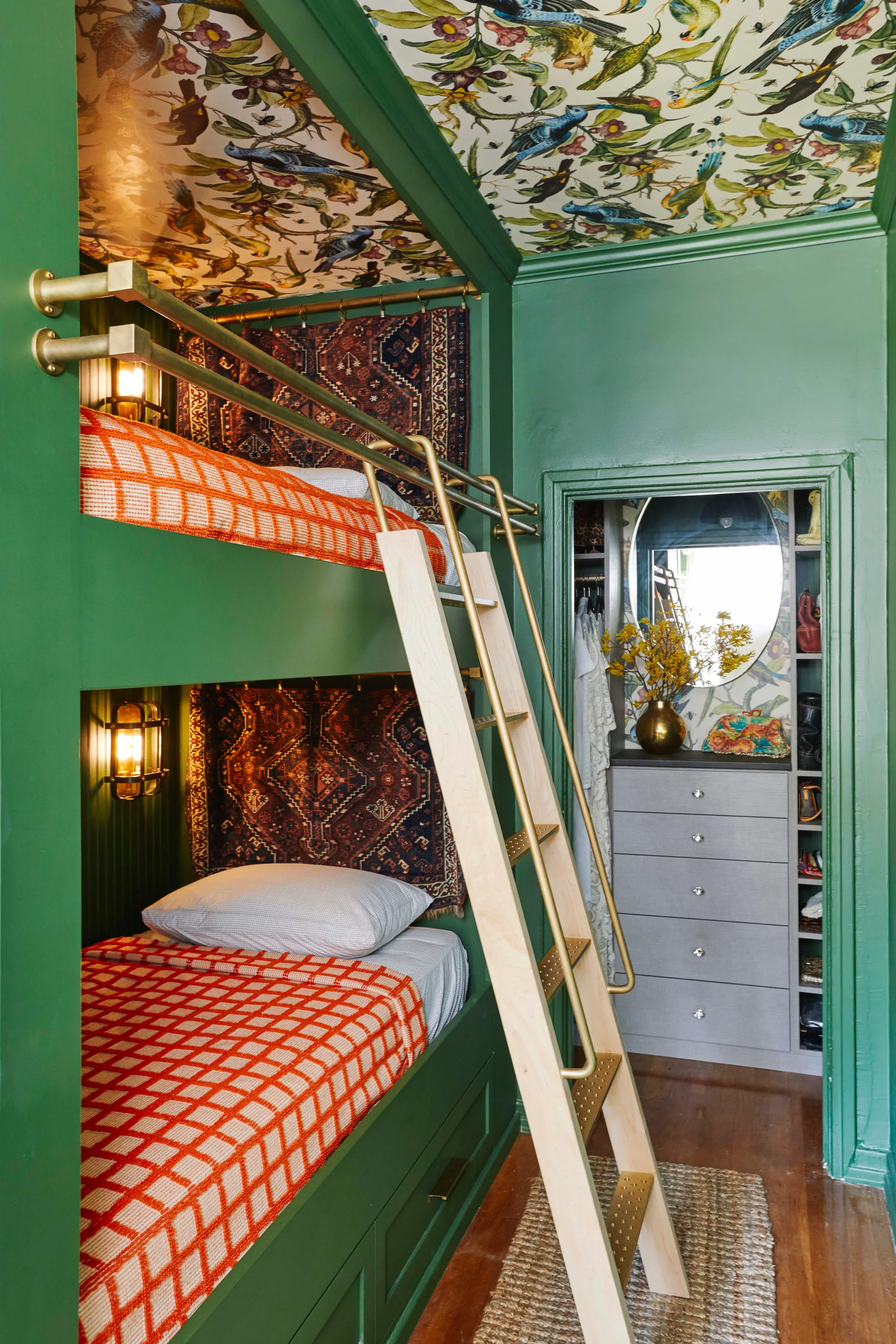 A green built-in bunk bed unit with orange blankets and a wooden ladder sits beneath a floral-patterned ceiling next to a walk-in closet.
