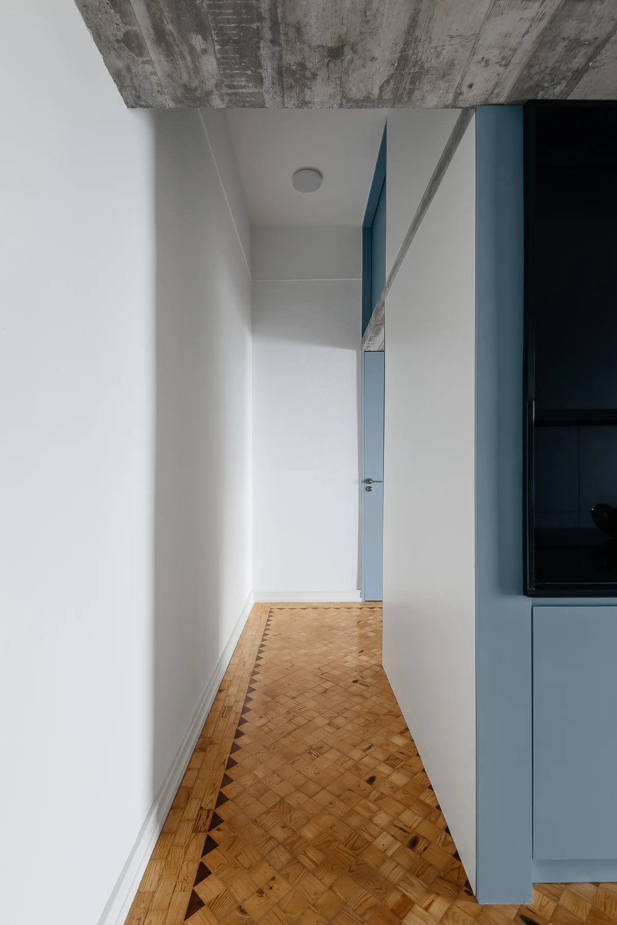 A narrow modern hallway features patterned basketweave parquet wood floors, white walls, an exposed concrete ceiling, and a light blue cabinet.