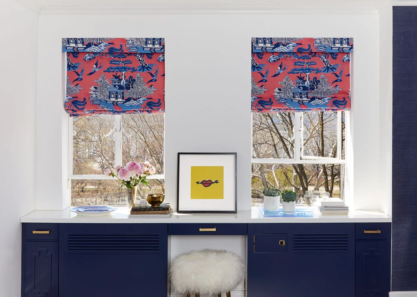 A built-in navy blue desk unit sits below two windows adorned with red and blue patterned Chinoiserie roman shades and a white fluffy stool.