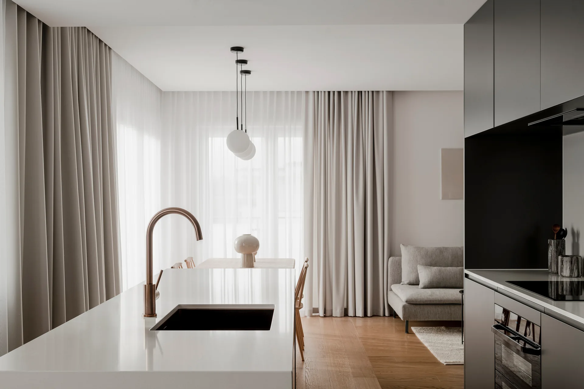 A minimalist kitchen featuring a white island with a copper faucet, dark grey cabinetry, and a dining area with pendant lights in the background.