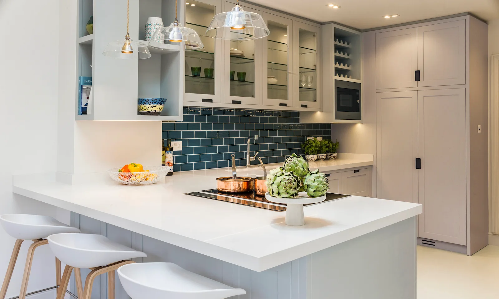 A modern kitchen featuring a light grey island with white barstools, glass pendant lights, and a dark teal subway tile backsplash.