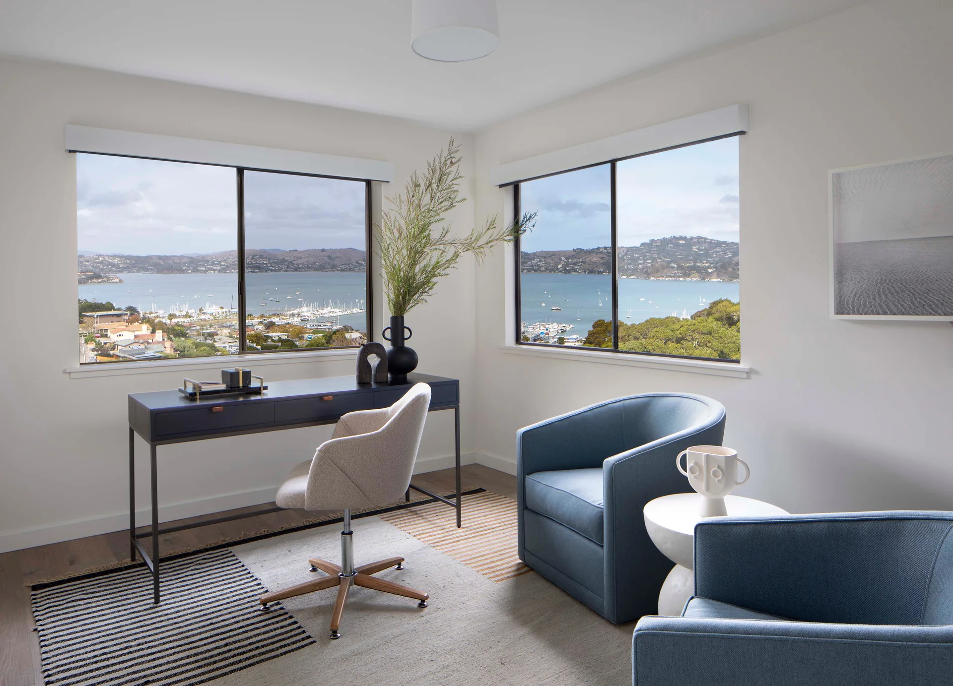 A modern home office features a dark writing desk, an upholstered swivel chair, two blue tub armchairs, and windows overlooking a harbor view.