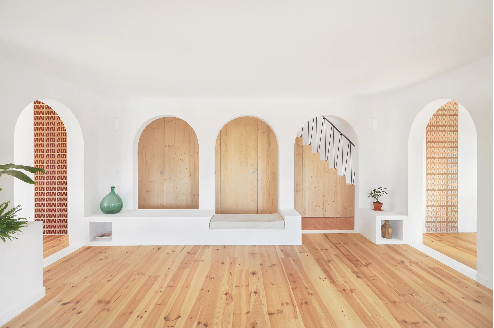 Multiple white plaster arches frame built-in light wood storage cabinets, a bench, a staircase, and geometric terracotta screens over a wood floor.