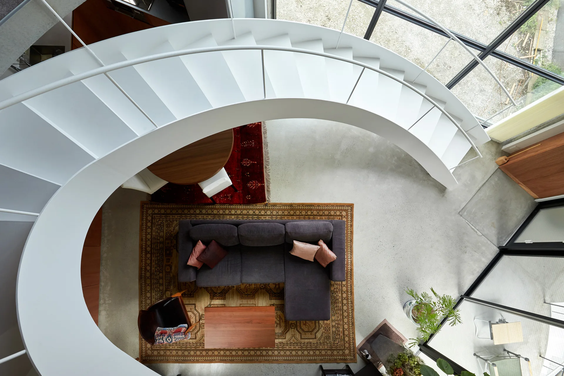 Overhead view of a living space with a white spiral staircase, charcoal sectional sofa, wooden coffee table, and patterned rugs on concrete.