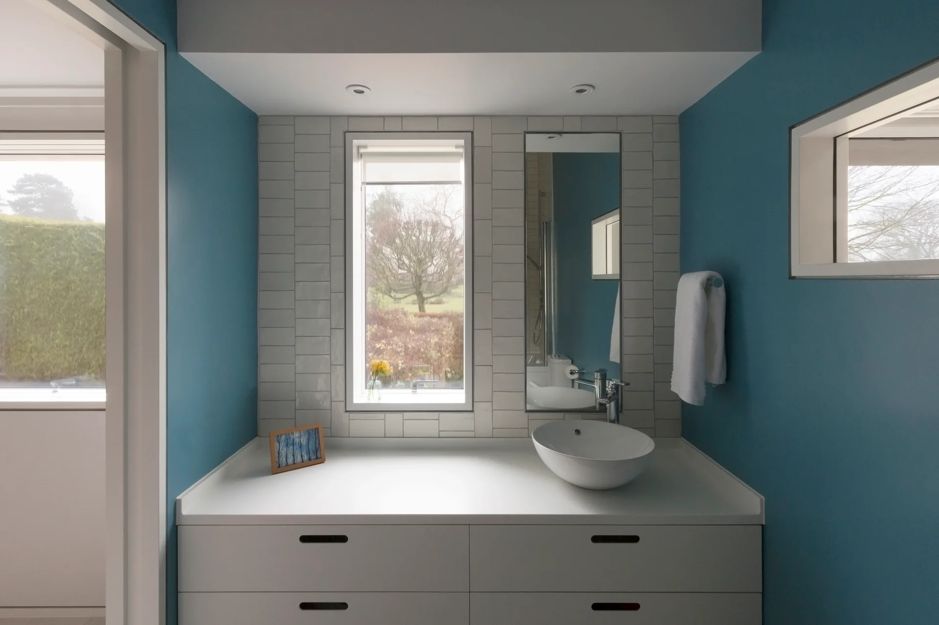 A modern bathroom vanity area with a white vessel sink, a vertical window, a matching mirror, and a blue wall accent.