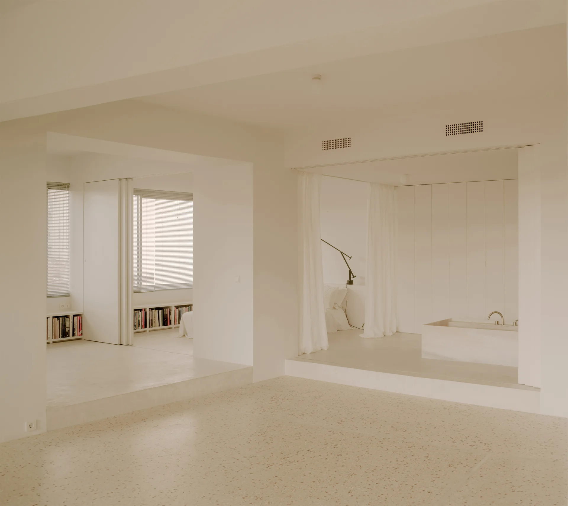 A minimalist open-plan bedroom features raised platforms, a built-in stone bathtub, white curtain partitions, and speckled terrazzo flooring.