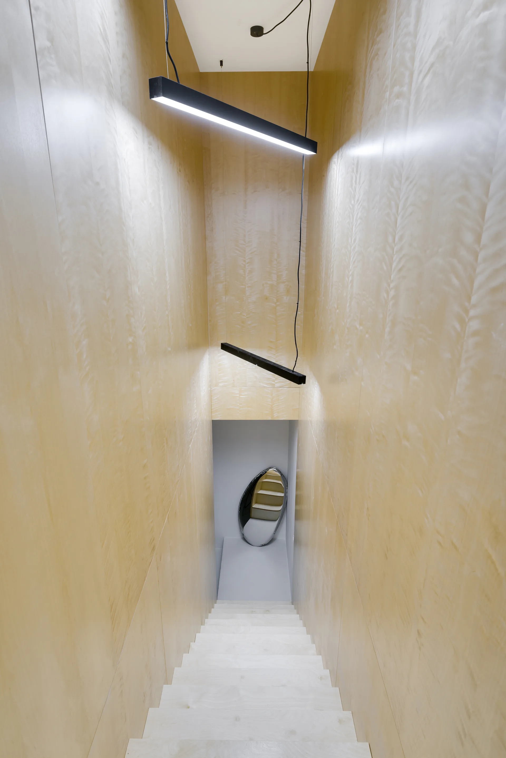 Looking down a minimalist staircase featuring light wood paneled walls, matching wooden steps, and angled black linear pendant lights.