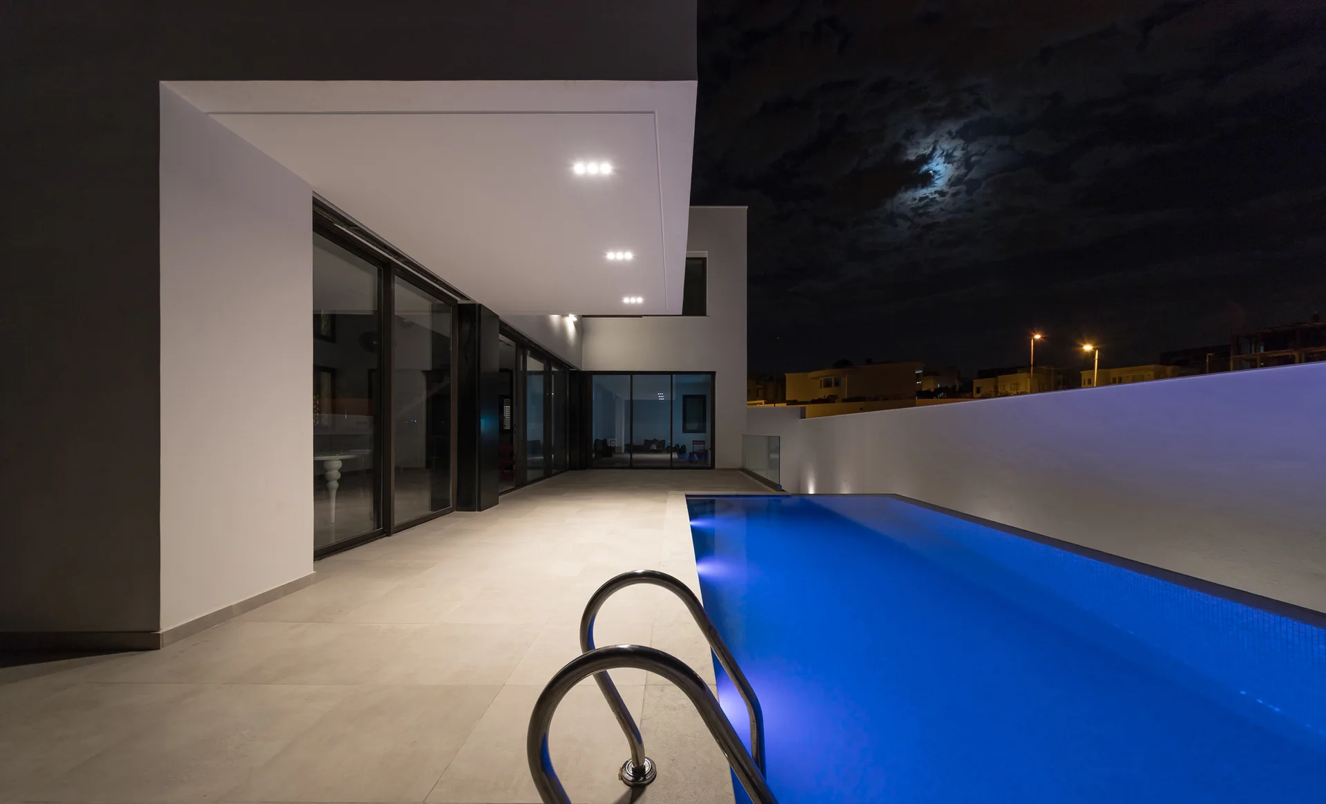 A minimalist patio at night features a glowing blue pool with a metal ladder next to white stucco walls and sliding glass doors.