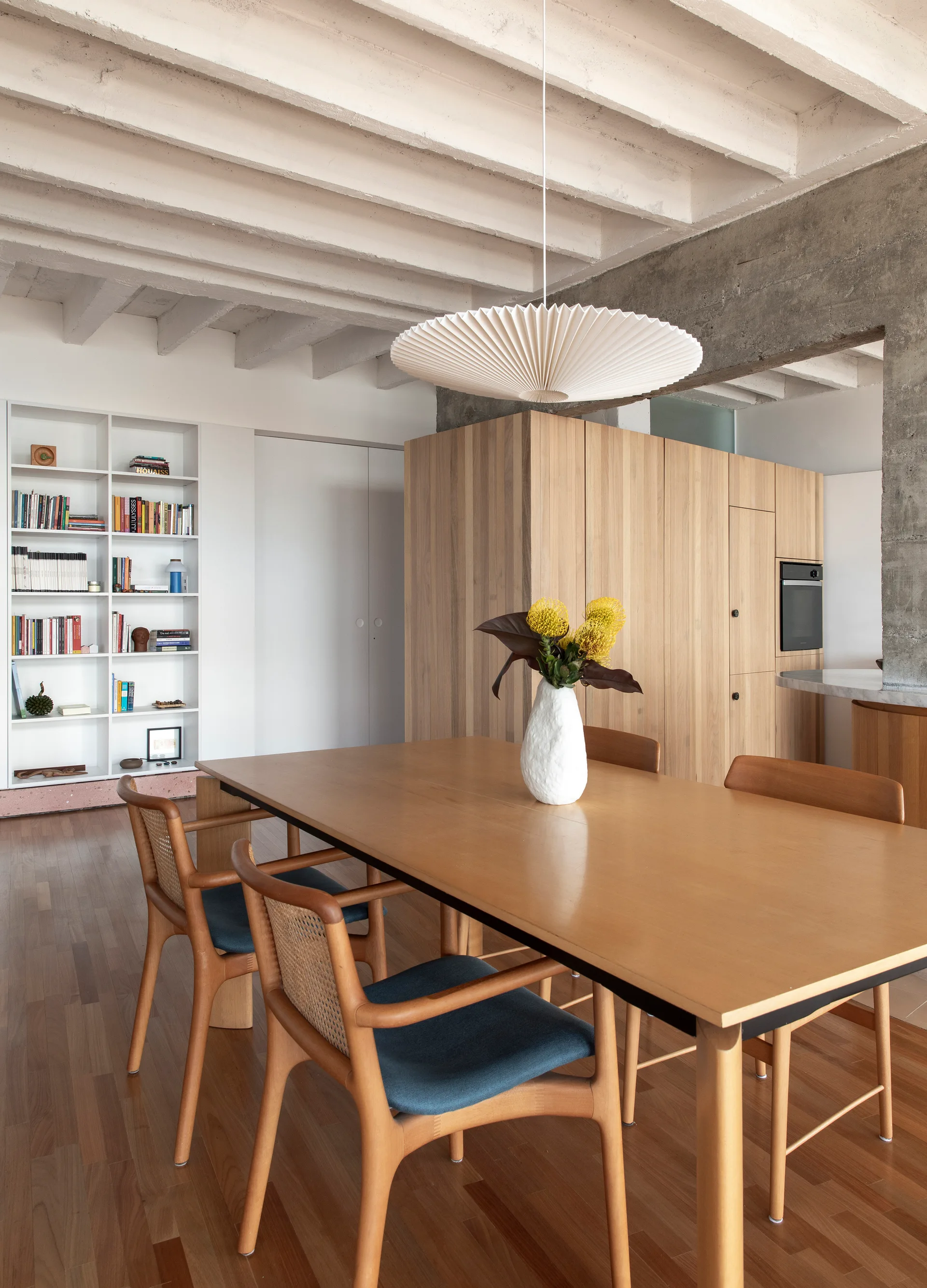 A wooden dining table and rattan chairs sit beneath a large pleated white pendant light, surrounded by wooden cabinets and white painted ceiling beams.