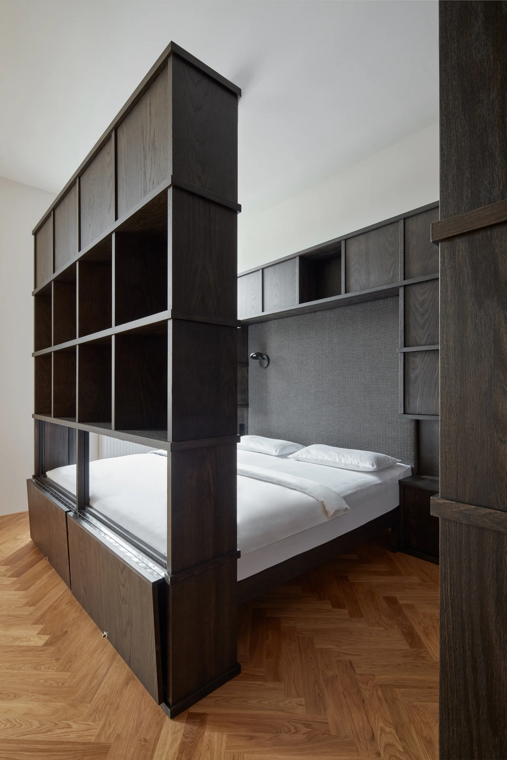 A dark wood room divider shelf stands next to a bed with white sheets and a grey patterned headboard on a herringbone wood floor.