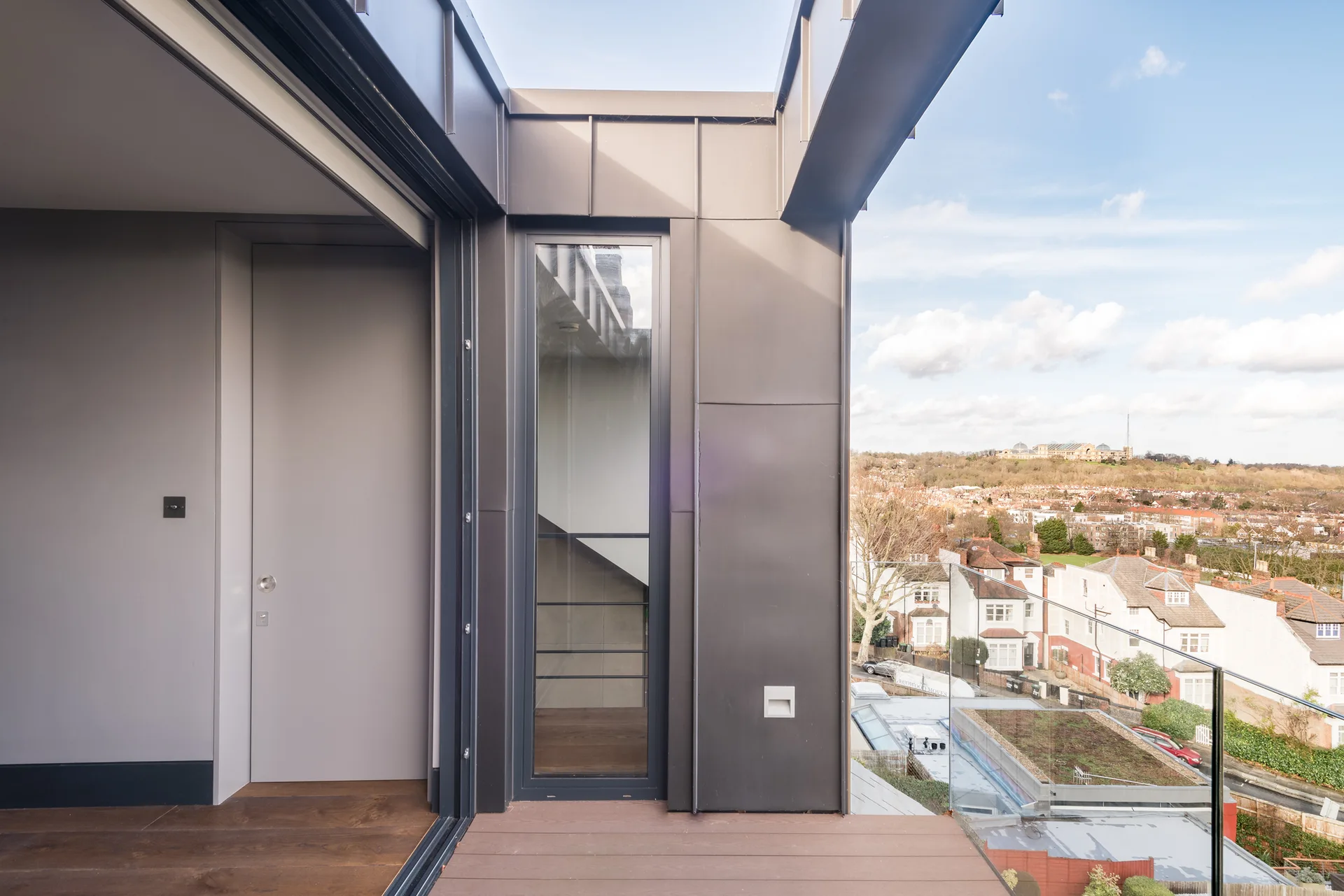 A modern balcony features dark grey metal cladding, wood-toned plank decking, and a clear glass railing overlooking a suburban neighborhood.