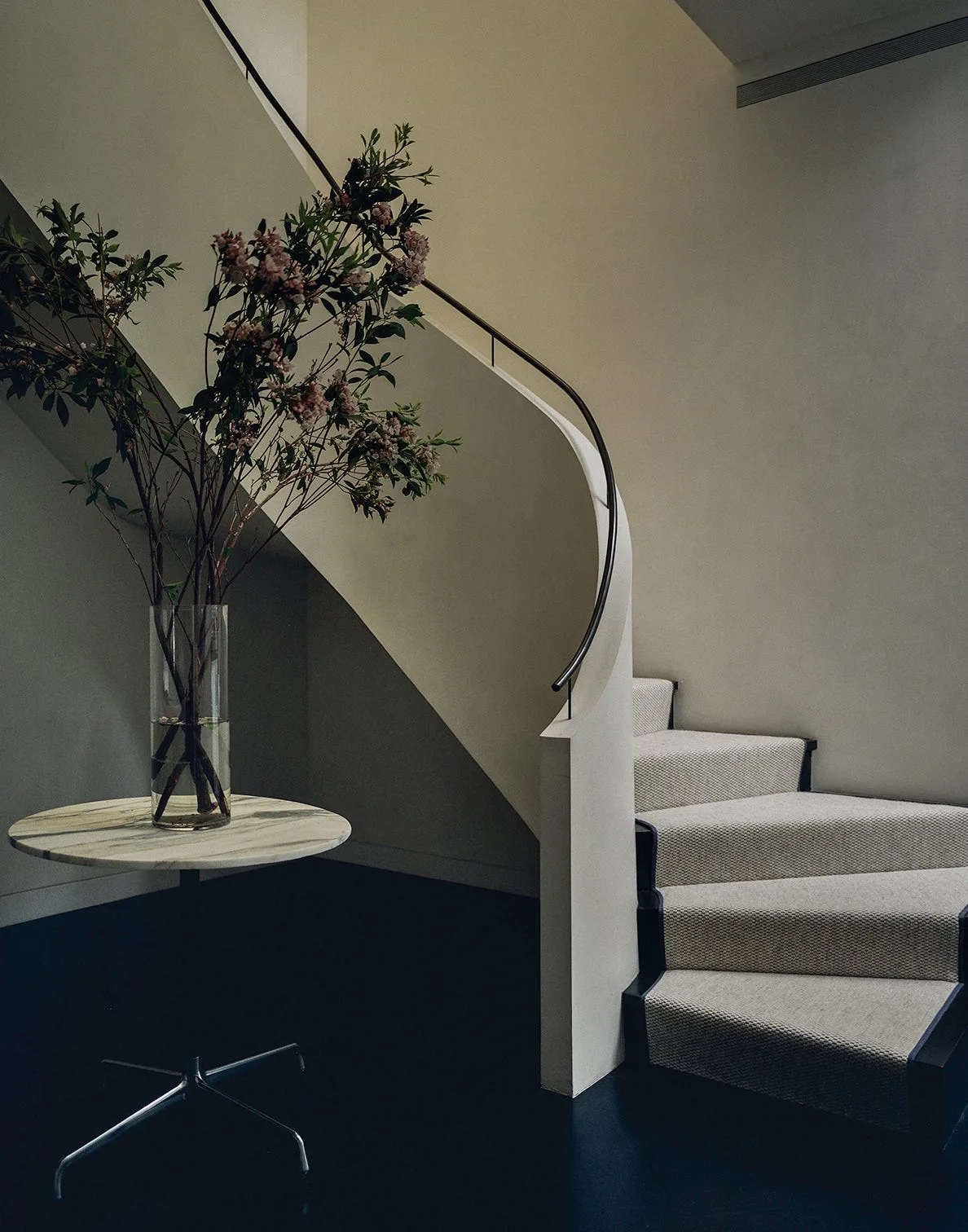 A curved staircase with a light woven runner and black trim stands next to a marble pedestal table holding a tall vase with branches.