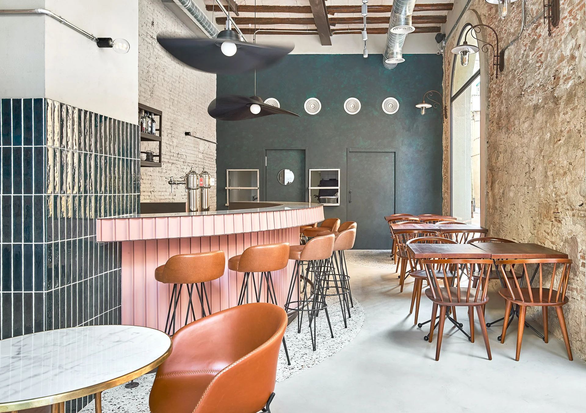 A contemporary restaurant interior features a curved pink fluted bar with brown leather stools alongside wooden dining tables and an exposed brick wall.