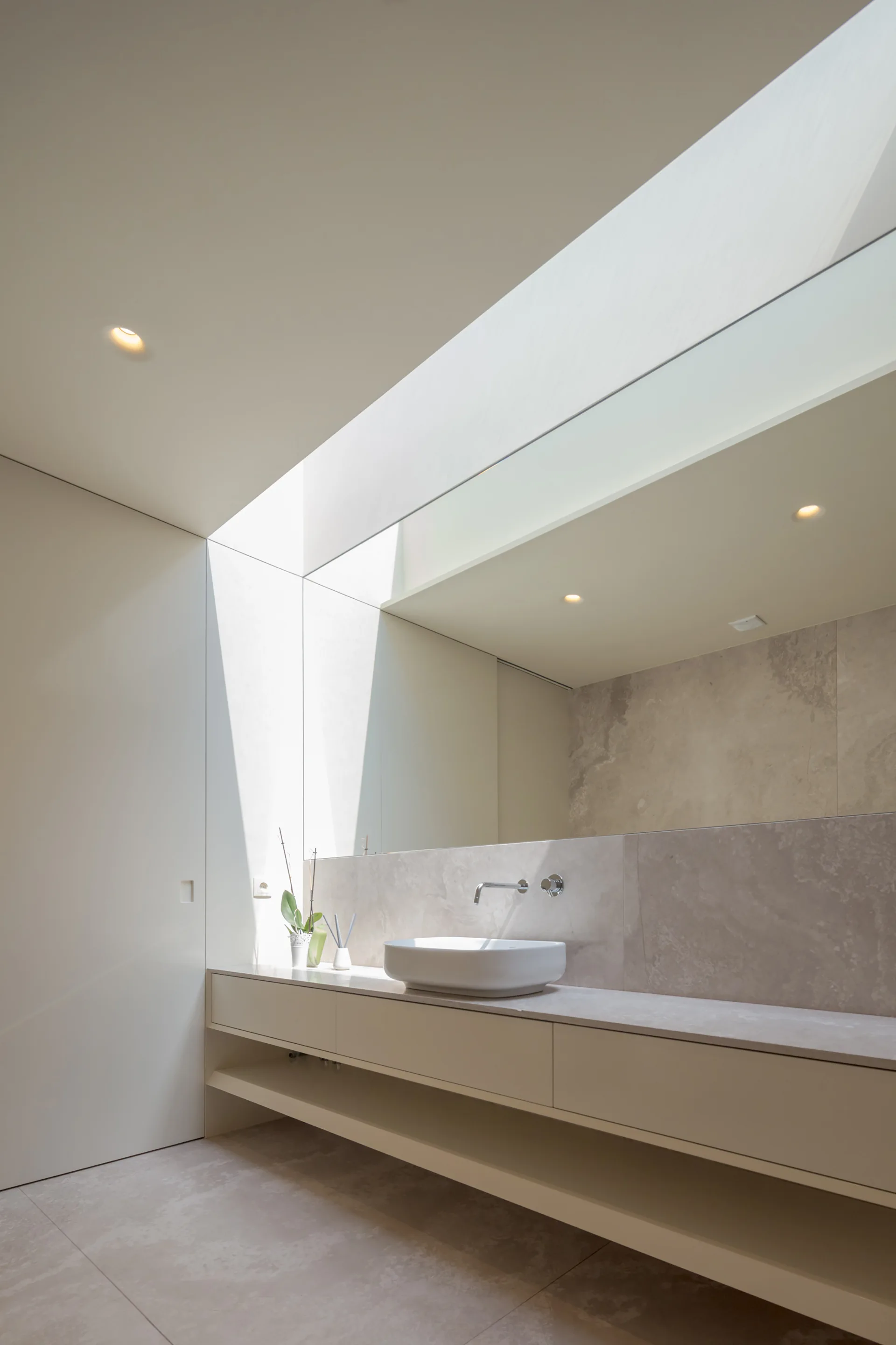A minimalist beige bathroom featuring a floating vanity with a vessel sink beneath a large skylight and a wall-mounted faucet.