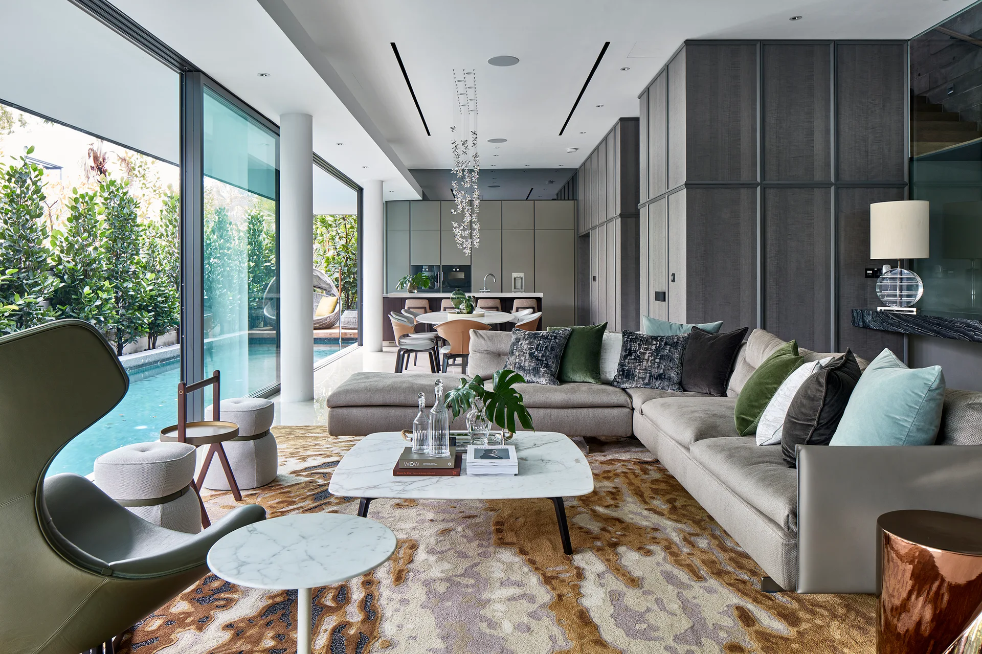 A modern open-plan living room features a grey sectional sofa, a marble coffee table, and sliding glass doors that open to a swimming pool.