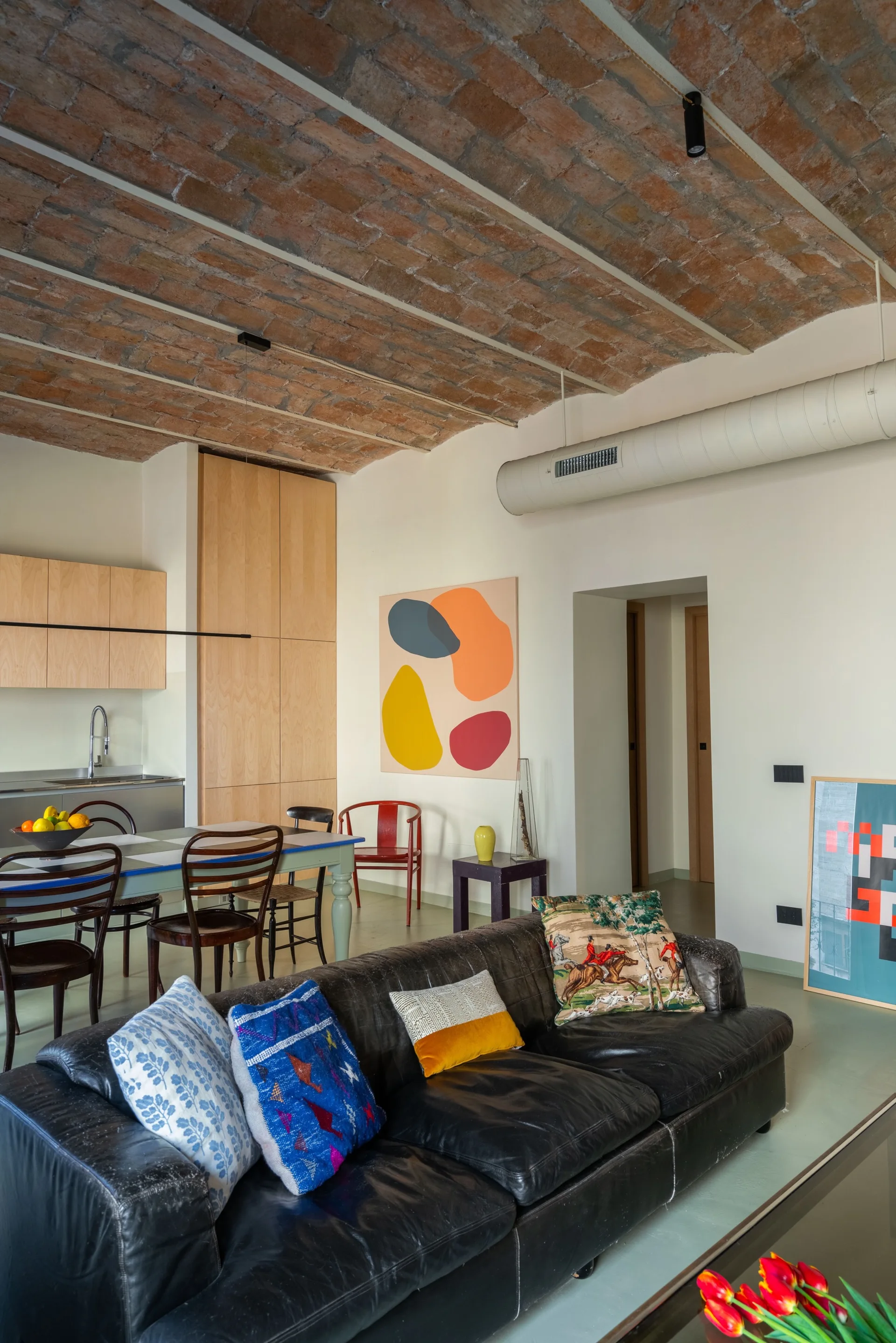 An open-concept living room features a black leather sofa with colorful pillows beneath a vaulted exposed brick ceiling and kitchen area.