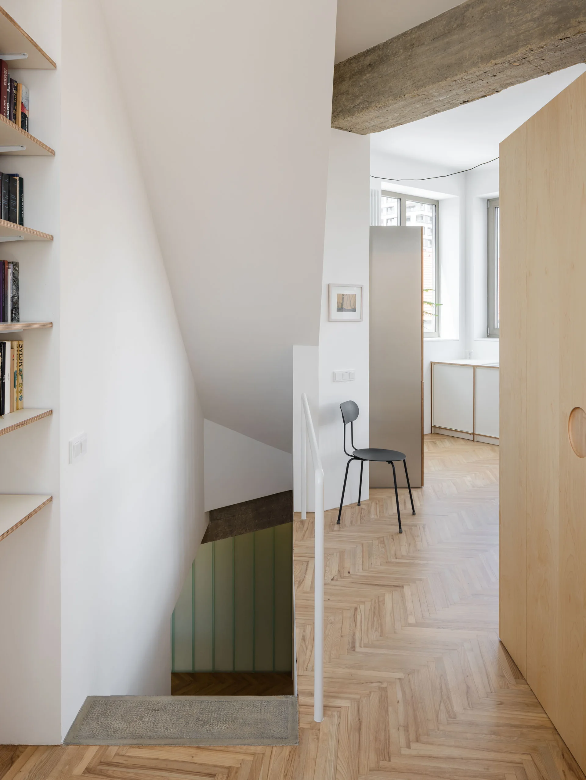 A minimalist hallway and staircase landing featuring light wood herringbone flooring, white walls, an exposed concrete ceiling beam, and a plywood door.