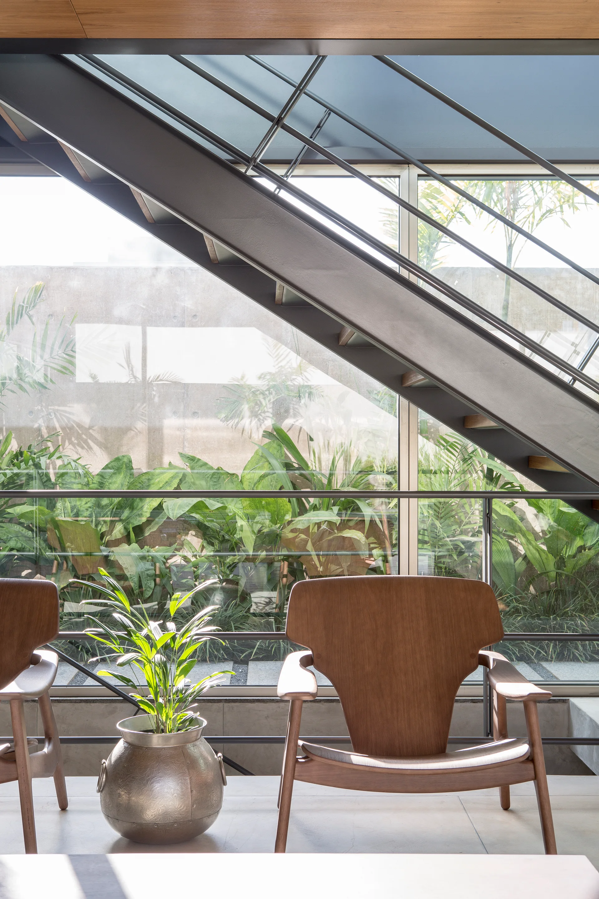Modern wood armchairs and a metallic planter sit before a large glass window overlooking a lush garden, with a metal staircase overhead.