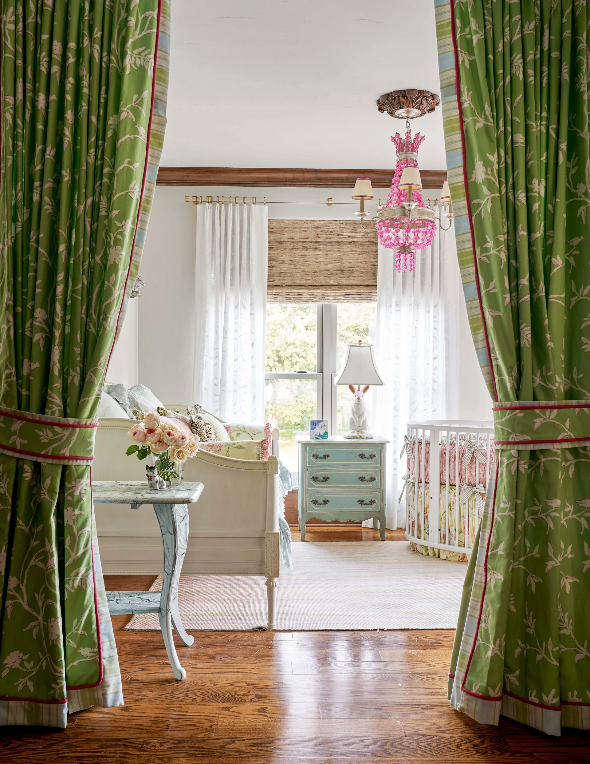 A whimsical nursery featuring a round white crib, a cream daybed, a distressed blue nightstand, and bold green floral curtains framing the room.