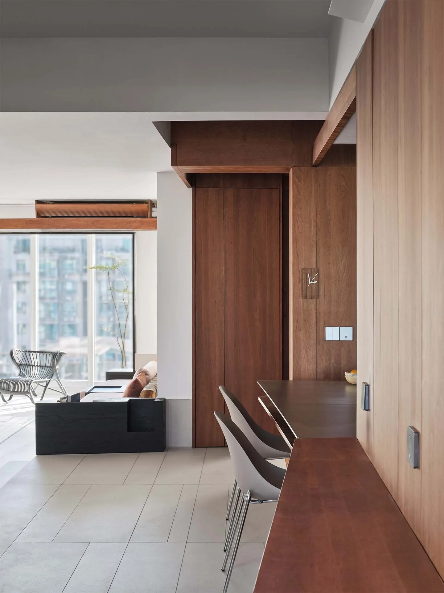 A modern apartment features warm wood wall paneling, a built-in kitchen counter with two gray stools, and a sunny living room in the background.