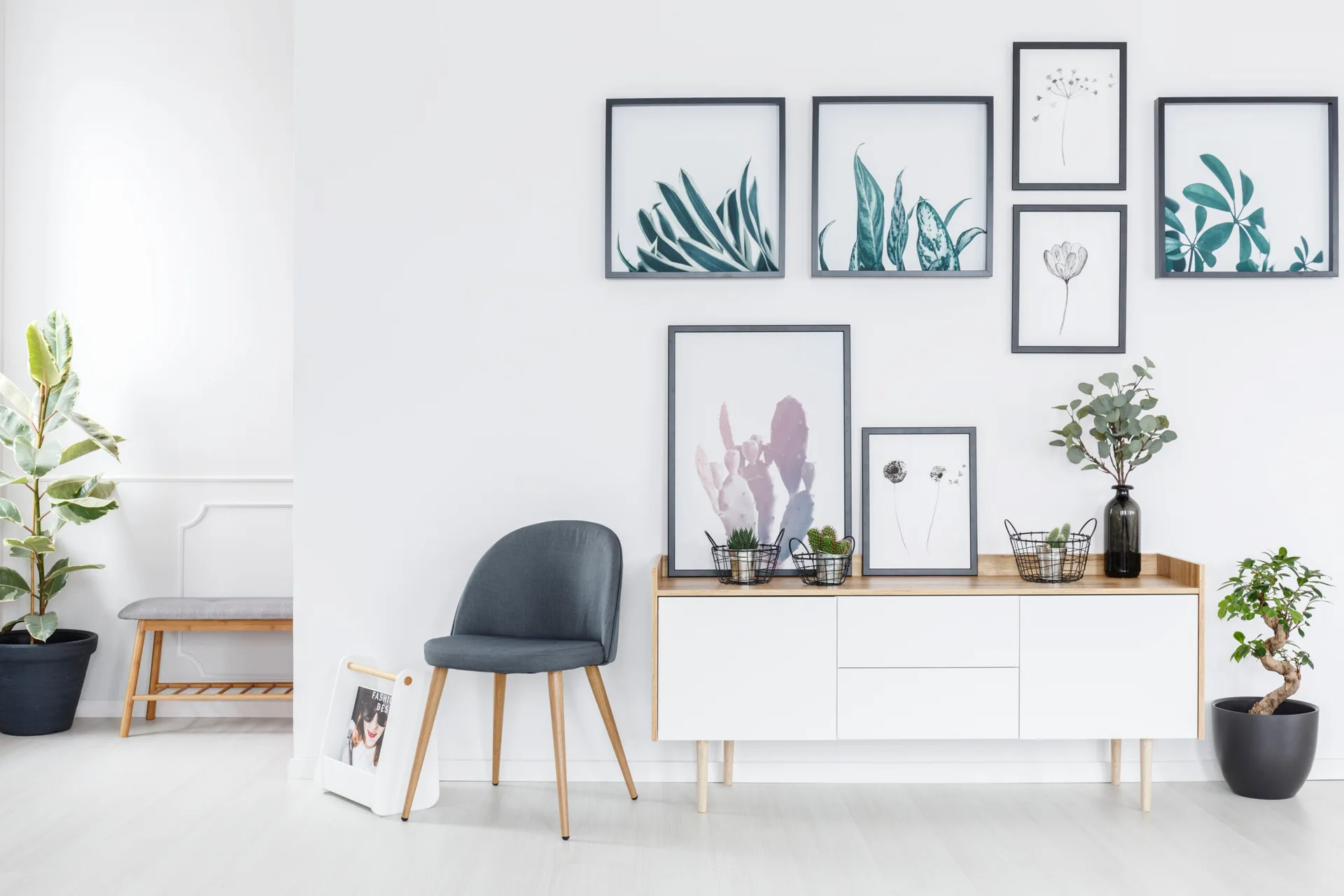 A white and wood sideboard sits beneath a gallery wall of framed botanical prints, next to a grey accent chair and potted plants.