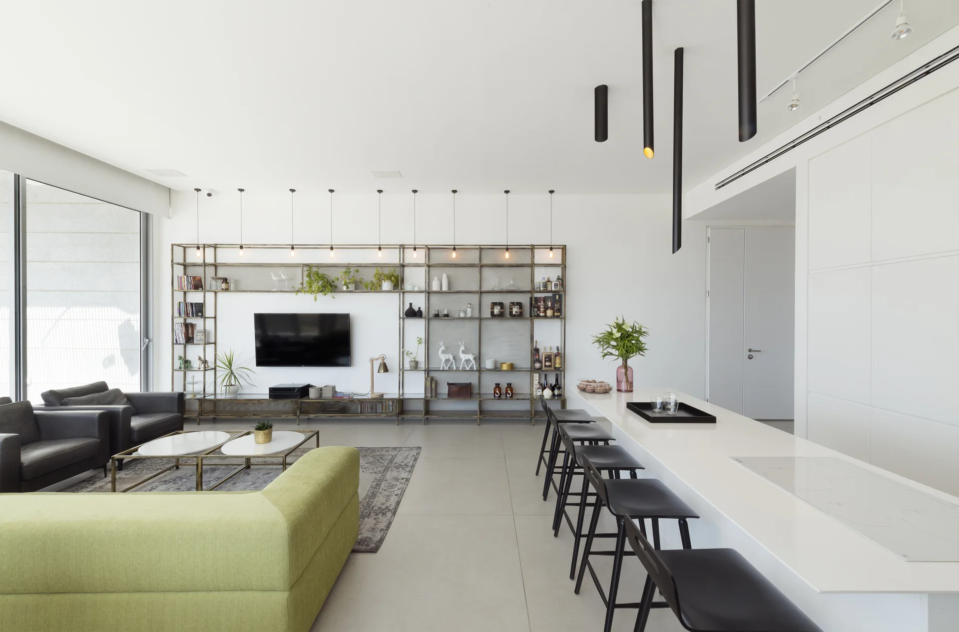 An open-plan living and kitchen area features a green sofa, a long white kitchen island with black bar stools, and a metal shelving unit on the wall.
