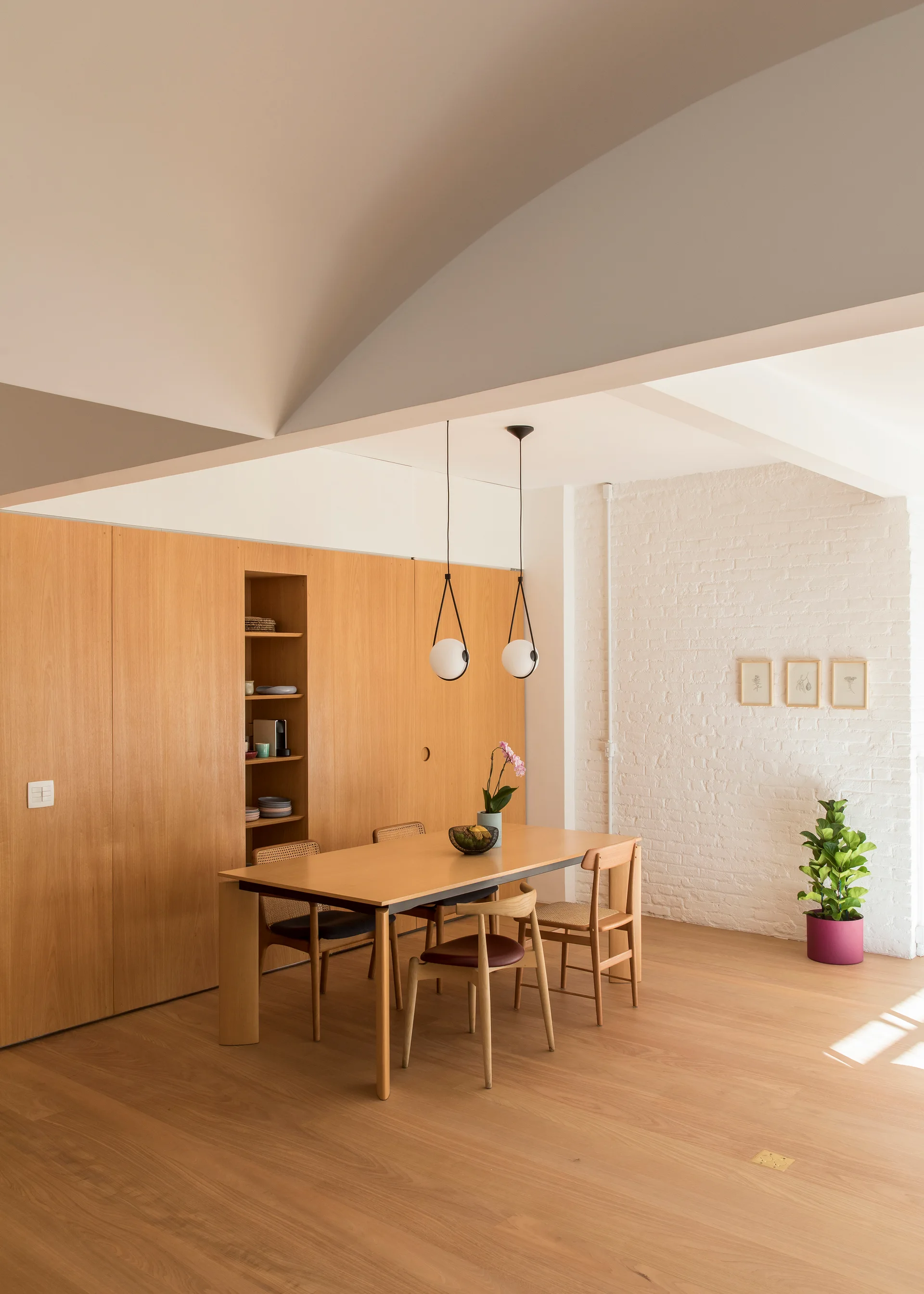 A minimalist dining room features a wooden table, cane-back chairs, spherical pendant lights, and a white brick wall next to seamless wood paneling.