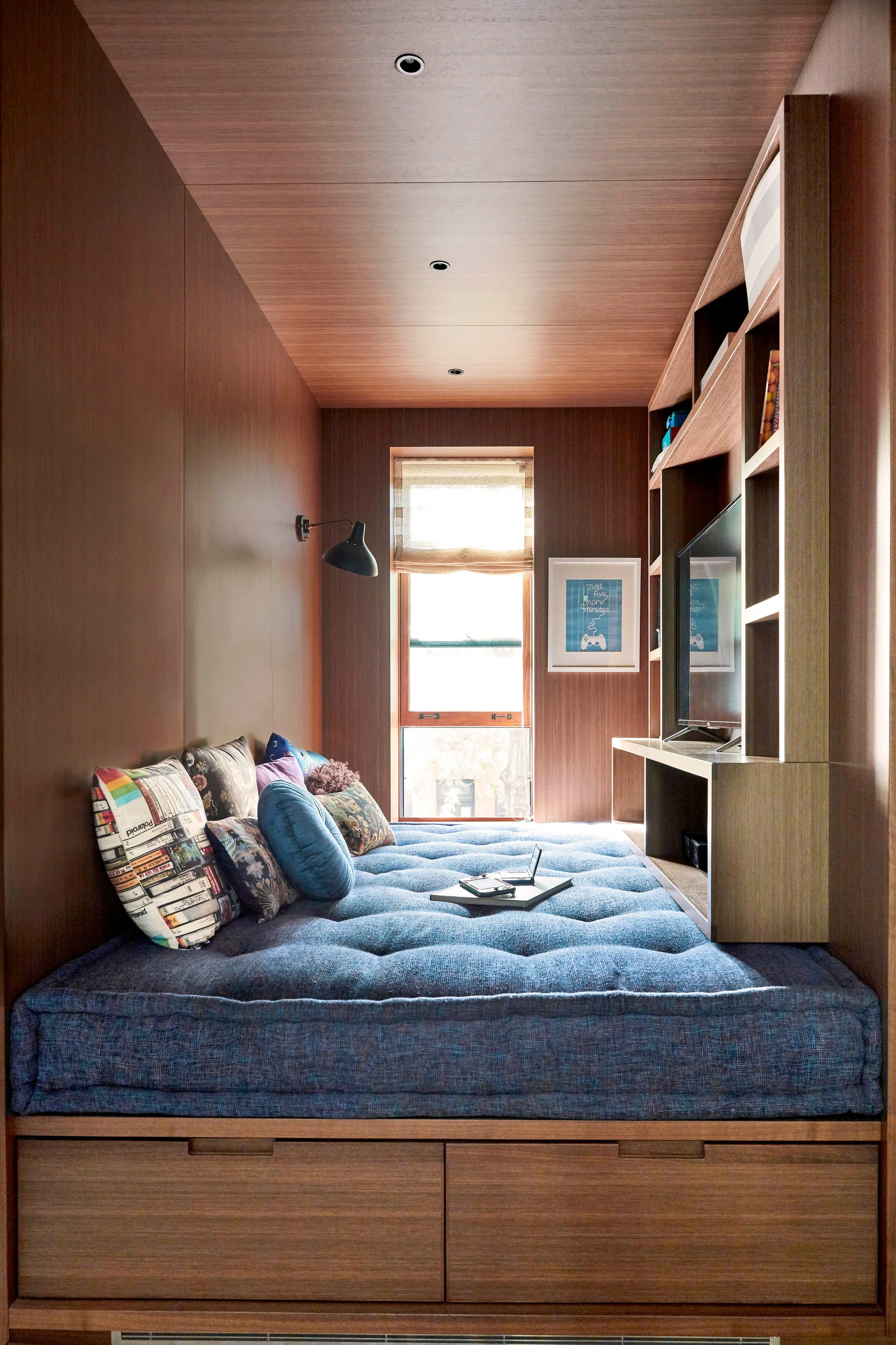 A compact wood-paneled sleeping nook features a built-in platform bed with a blue tufted mattress, storage drawers, and integrated shelving.