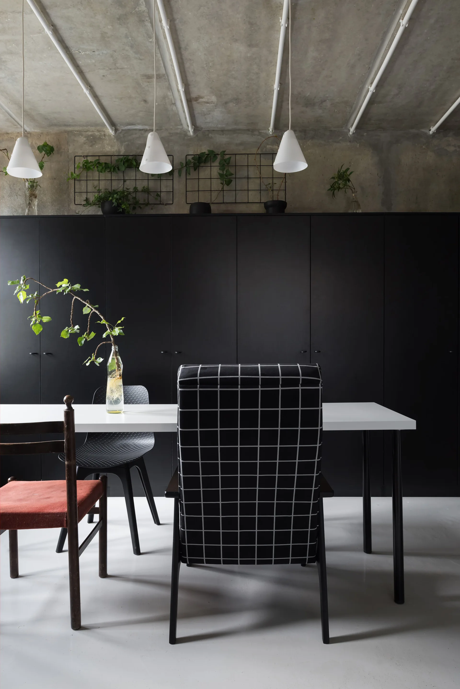 A modern dining area features a white table with mixed chairs set against a full wall of matte black cabinets and an exposed concrete ceiling.
