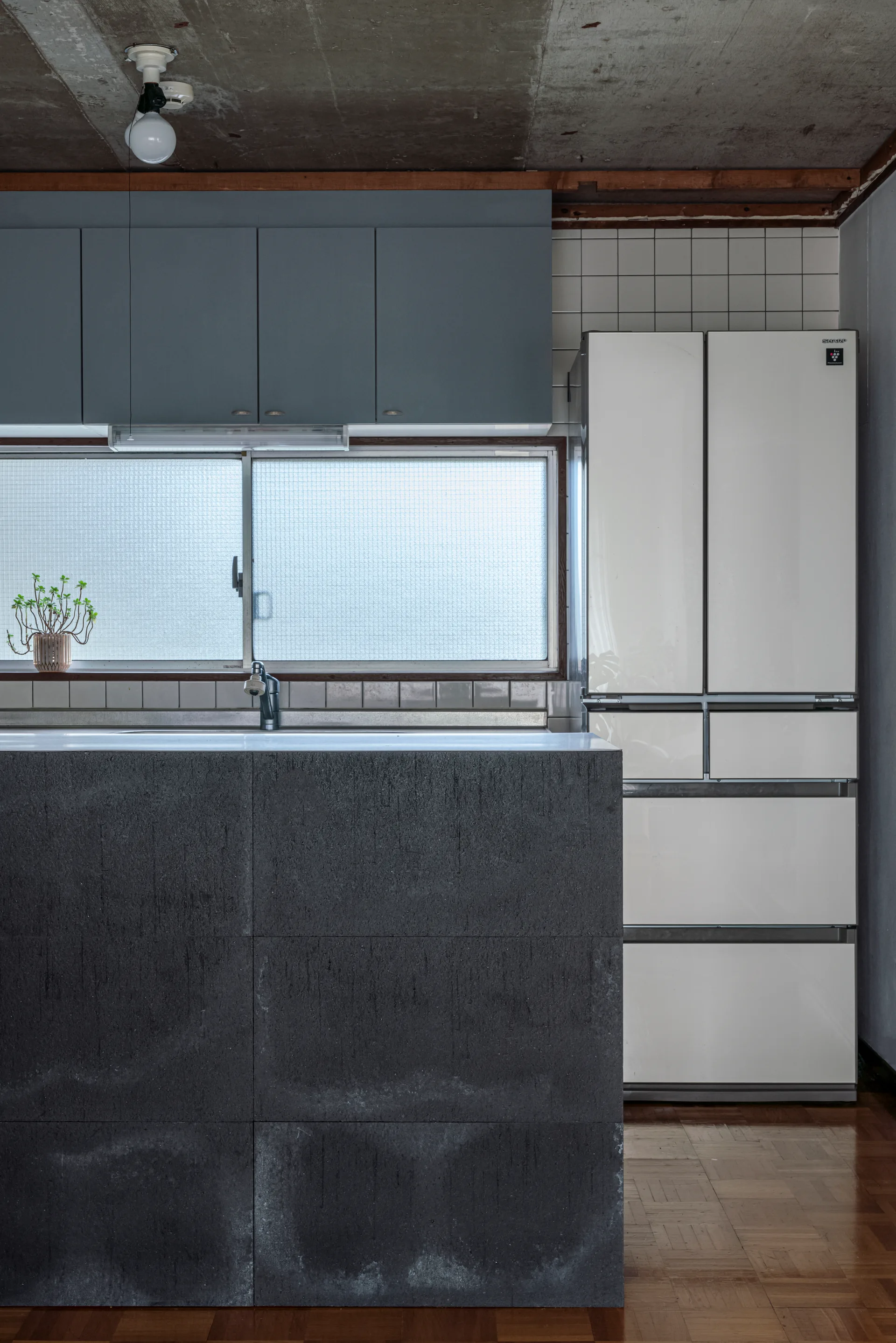 A minimalist kitchen features a textured dark stone island, matte grey cabinets, a white refrigerator, and an exposed raw concrete ceiling.