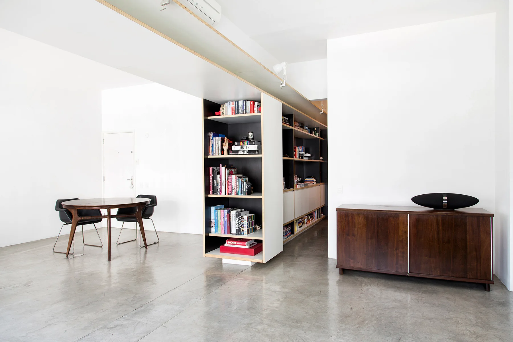 A minimalist open-plan room features a large double-sided wooden bookshelf divider, a round dining table, a wooden sideboard, and polished concrete floors.