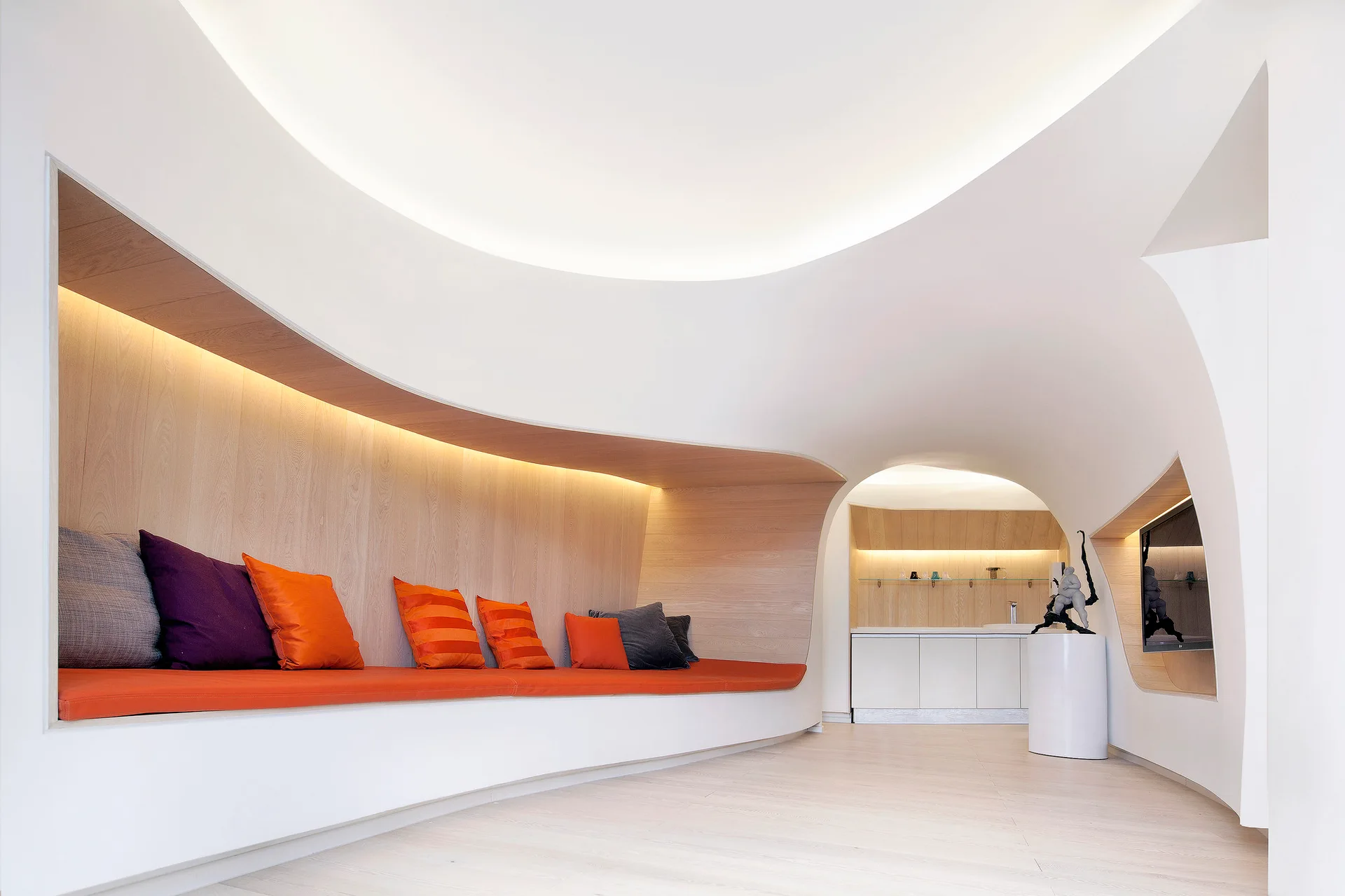 A minimalist white room featuring a curved built-in bench with orange cushions set inside a recessed light wood wall niche under integrated lighting.