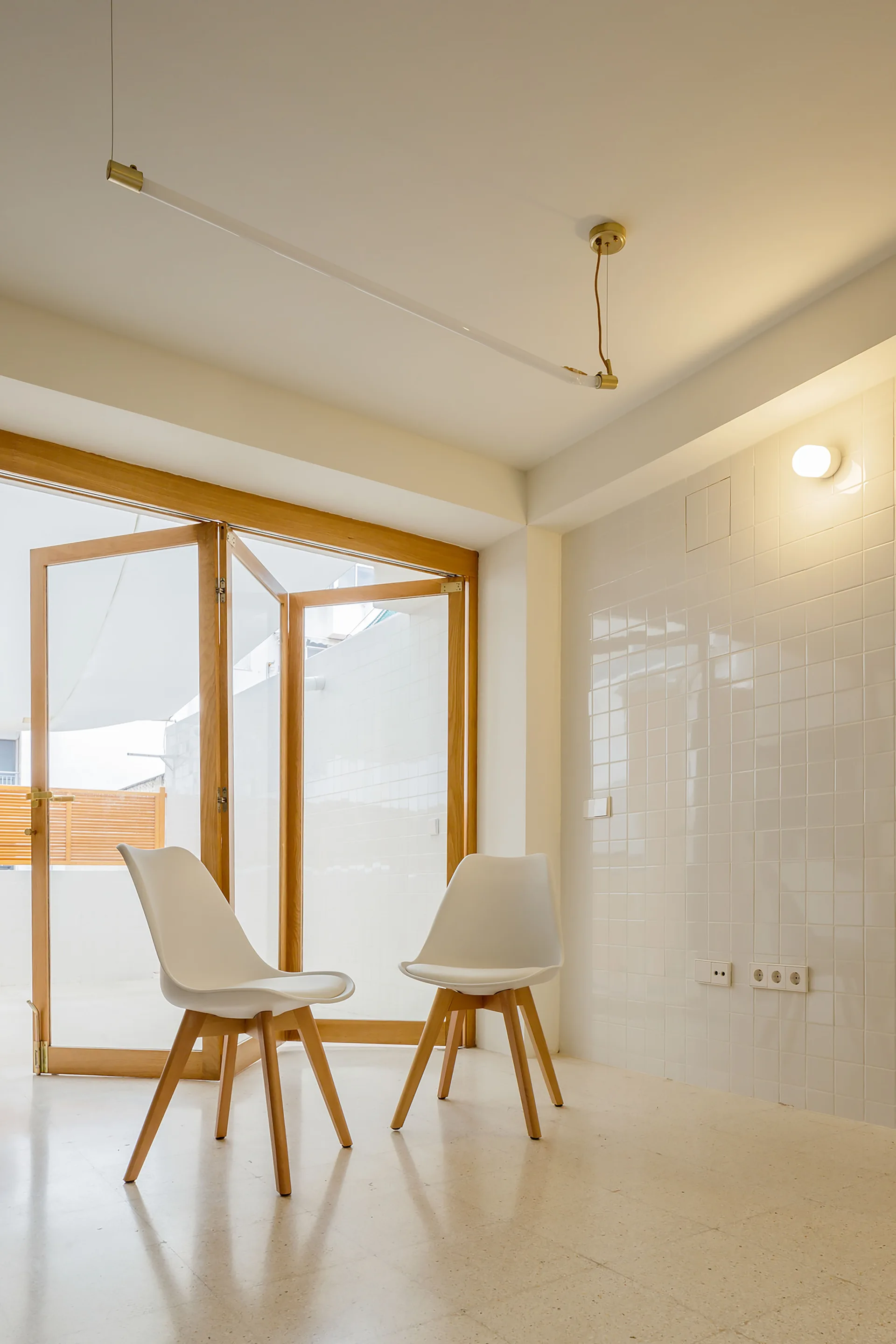 Two white modern chairs sit on a speckled floor next to a glossy white tiled wall and a large wood-framed glass folding door.
