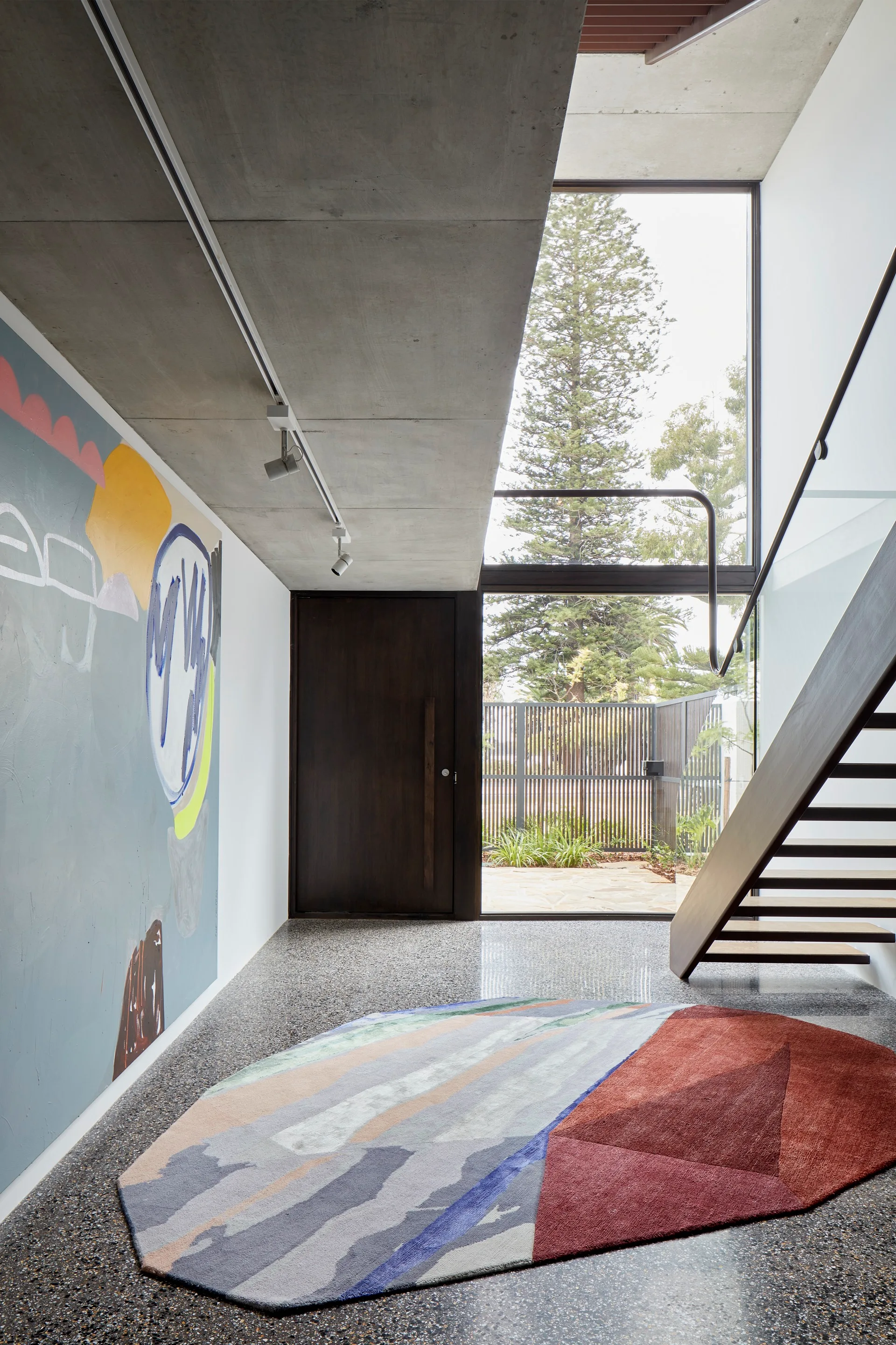 A modern entry foyer featuring a polished terrazzo floor, an open riser wooden staircase, and an exposed concrete ceiling with a colorful abstract rug.