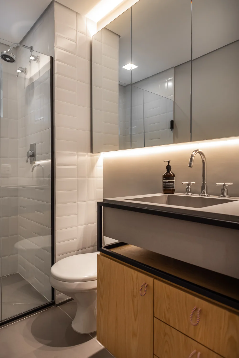 A modern bathroom features a wood vanity with a gray concrete sink, a glowing mirror cabinet, a toilet, and a glass-enclosed shower.