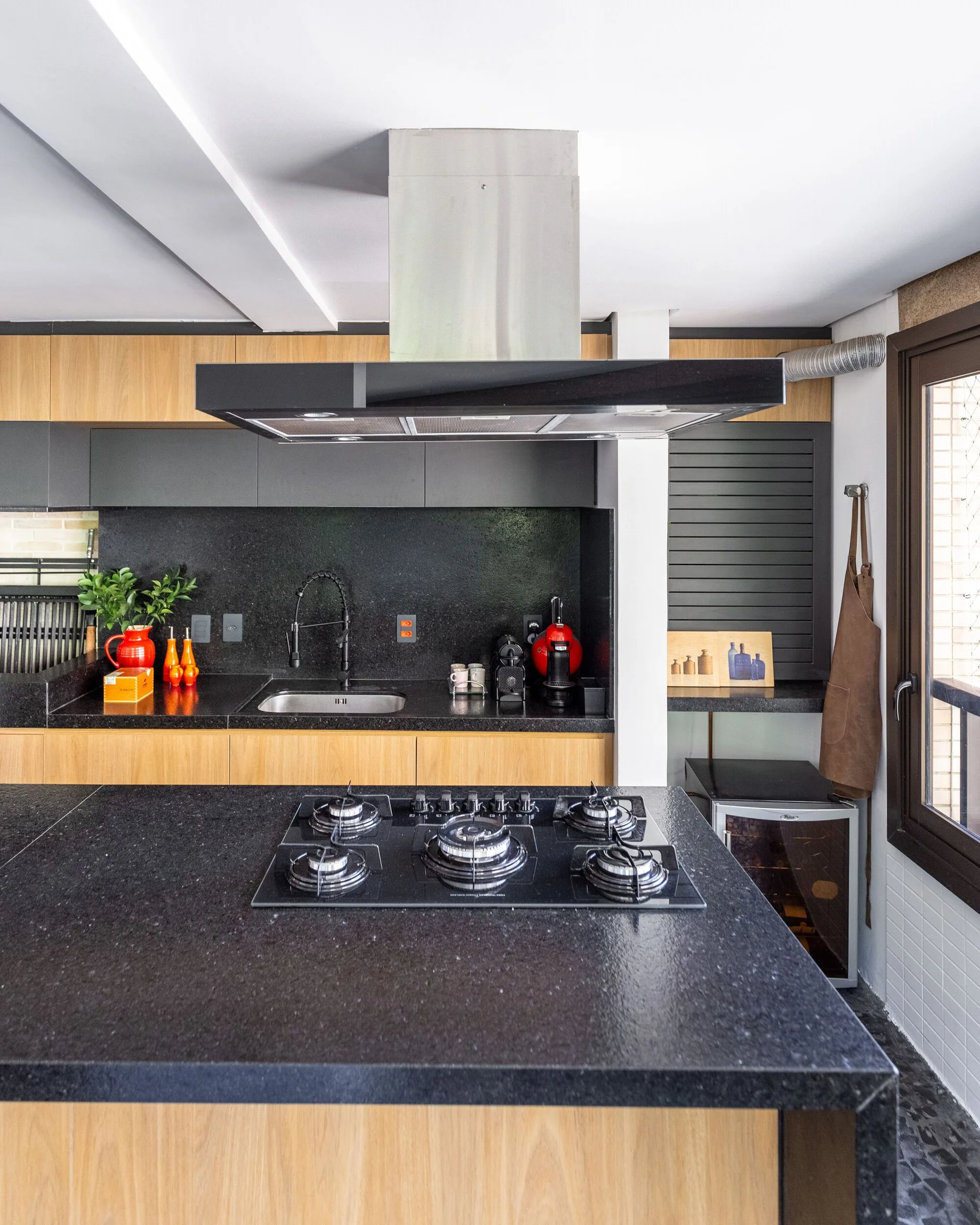 A contemporary kitchen features a black stone island with an integrated cooktop, wood and charcoal grey cabinetry, and a black undermount sink.
