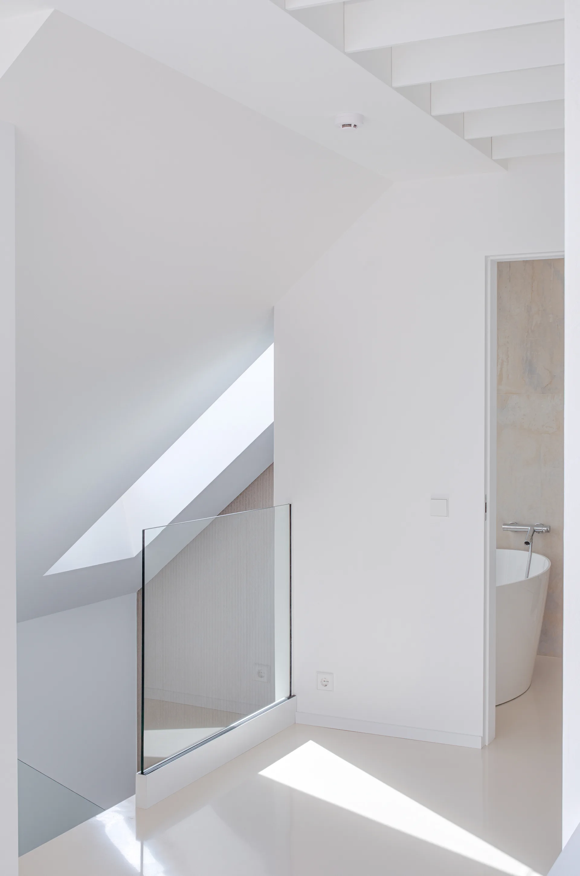 A minimalist white landing features a frameless glass staircase railing, sloped ceiling with a skylight, and a doorway leading to a bathroom.