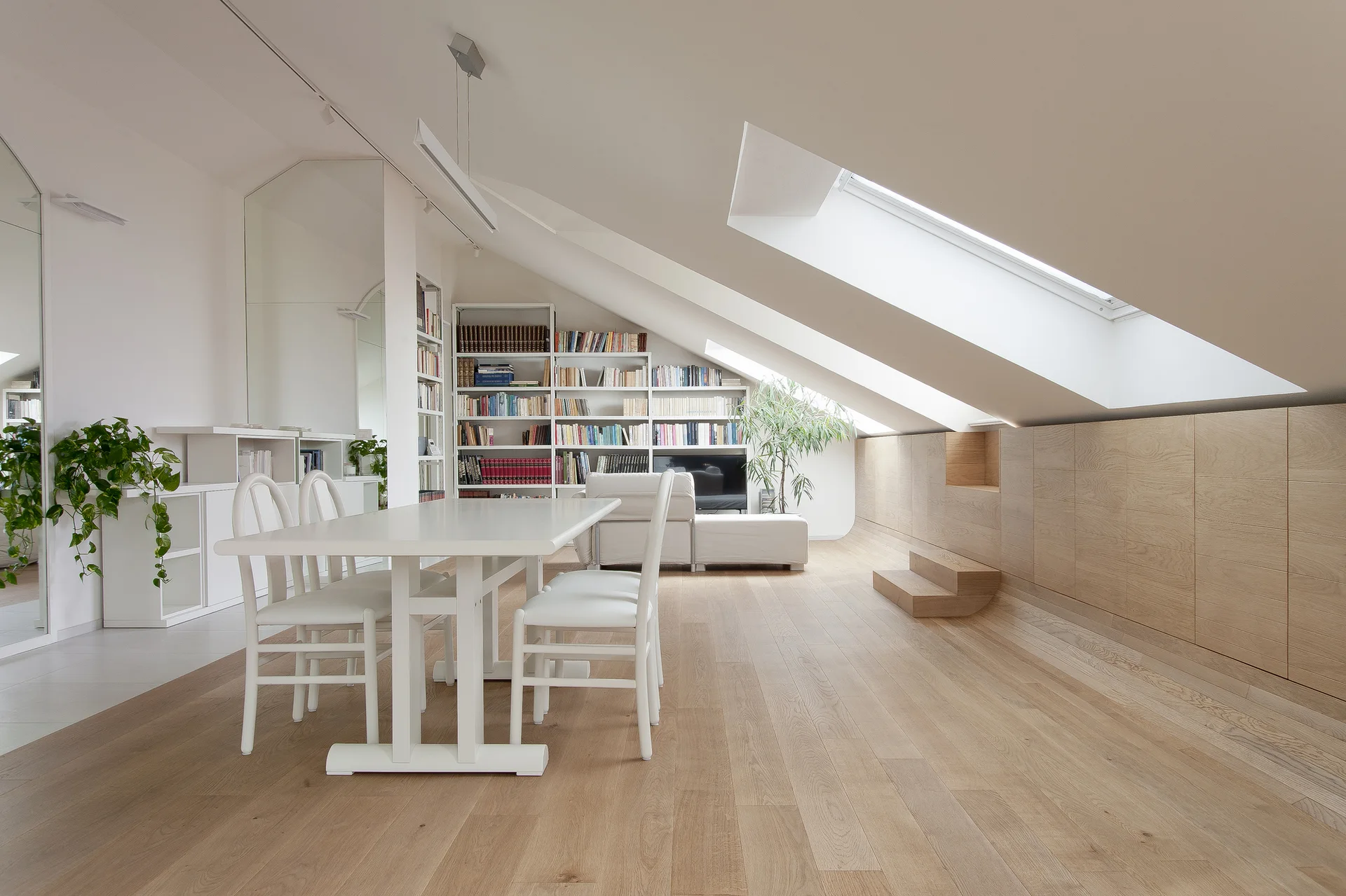 A bright attic dining room features a white table and chairs, light wood flooring, and sloped ceilings with multiple skylights.