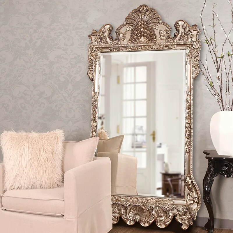 A large ornate floor mirror with a carved gold frame stands next to a beige armchair with a fluffy pillow against damask wallpaper.