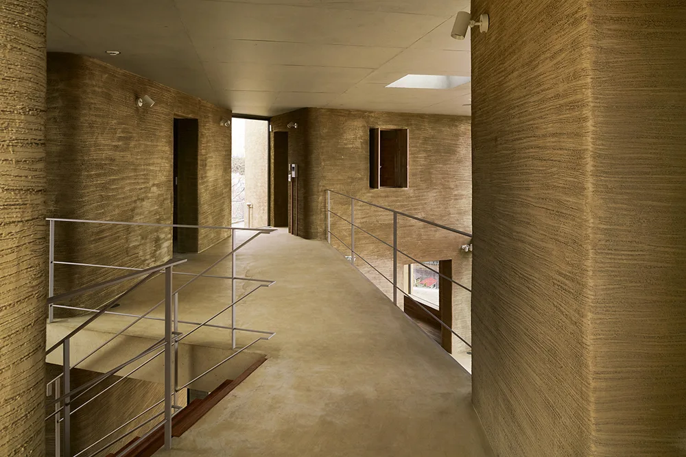 A minimalist hallway landing featuring rough textured ochre plaster walls, a thin metal staircase railing, and natural light from a skylight.