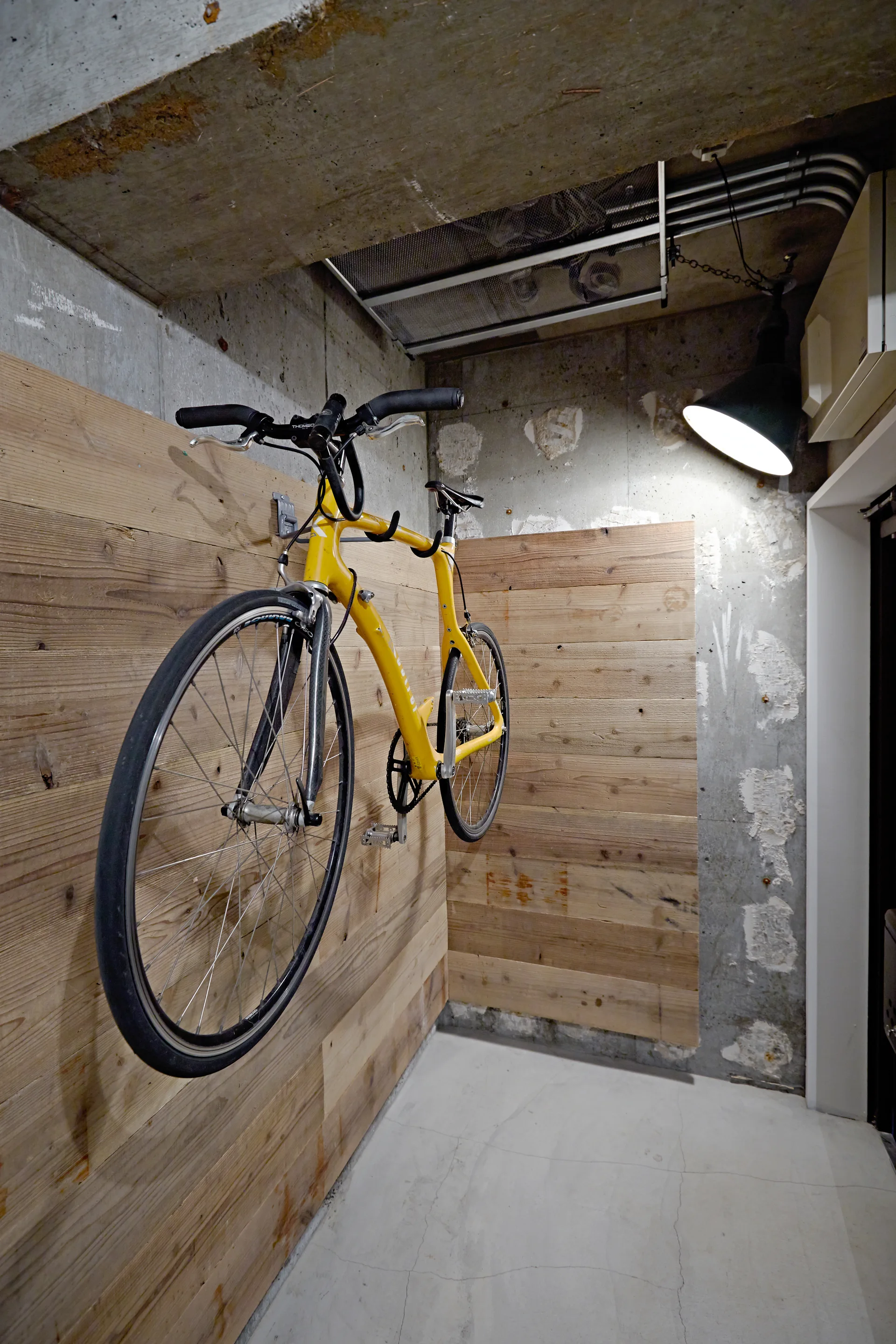 A bright yellow bicycle hangs on horizontal wood panels in an industrial corner with exposed concrete walls, concrete ceiling, and a black pendant light.