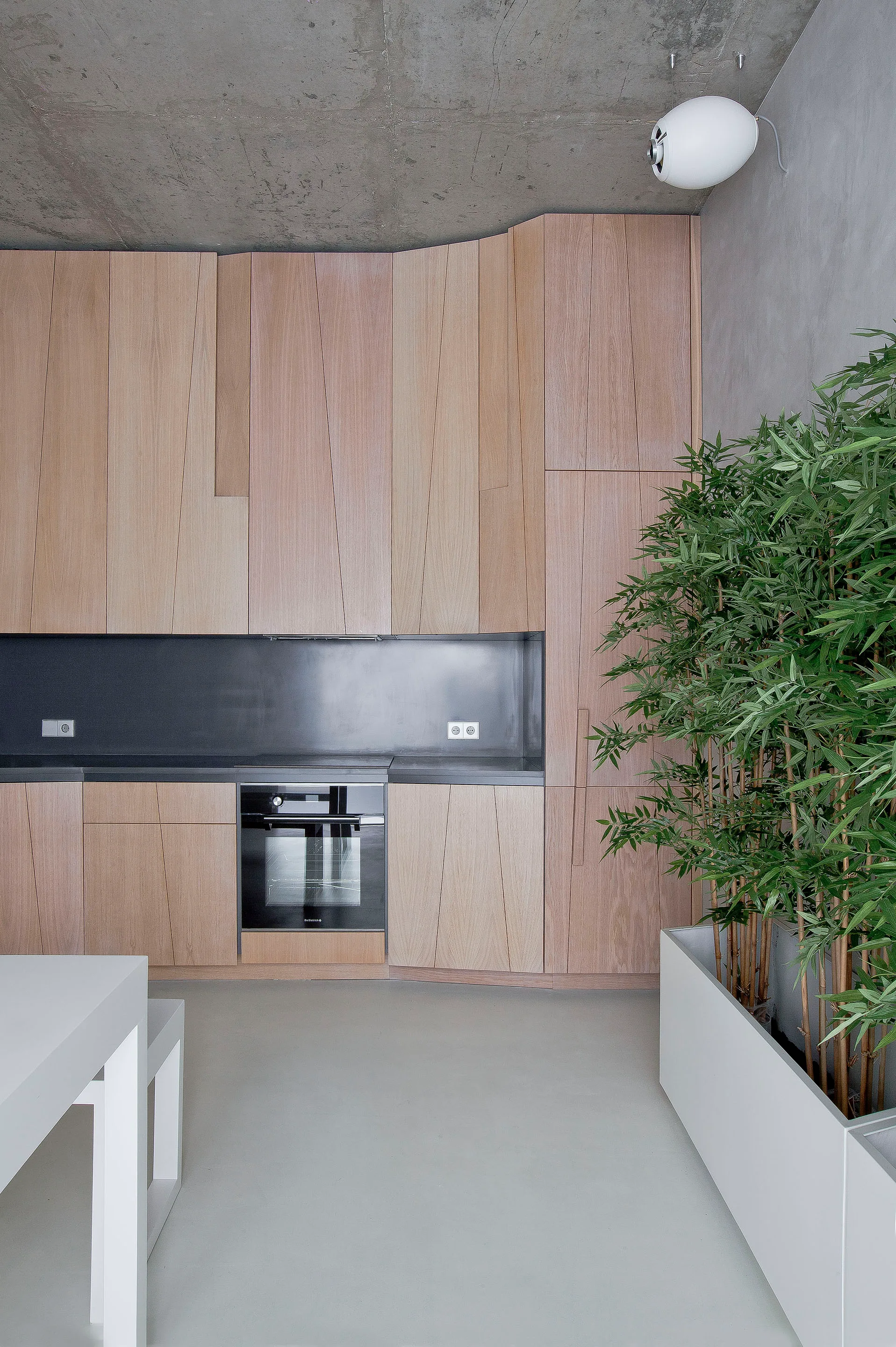 A modern kitchen features light wood cabinets with angled geometric paneling, an integrated black oven, a dark backsplash, and an exposed concrete ceiling.