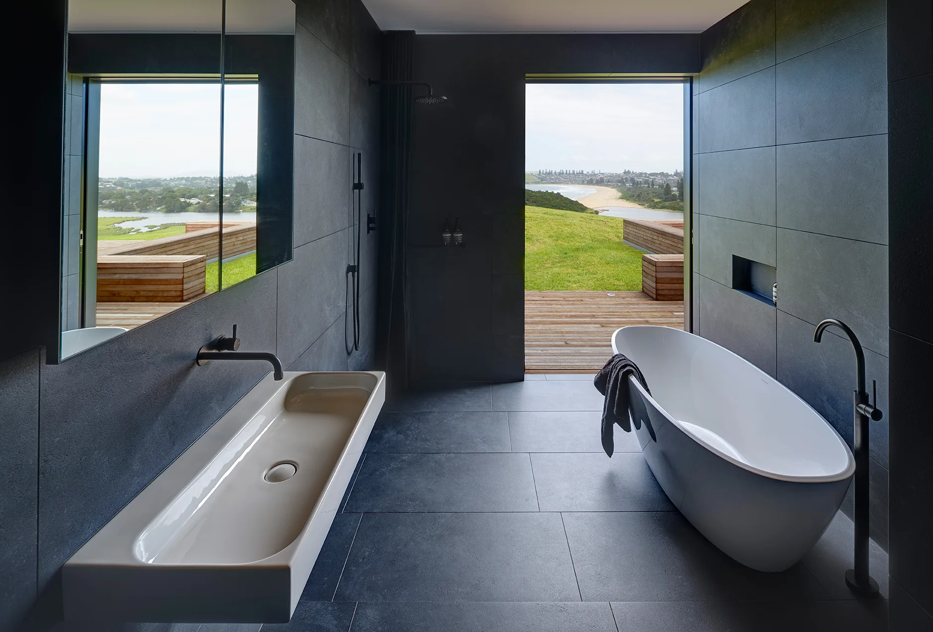 A modern charcoal tiled bathroom features a white freestanding bathtub, a long wall-mounted sink, and a large open doorway leading to a wooden deck.